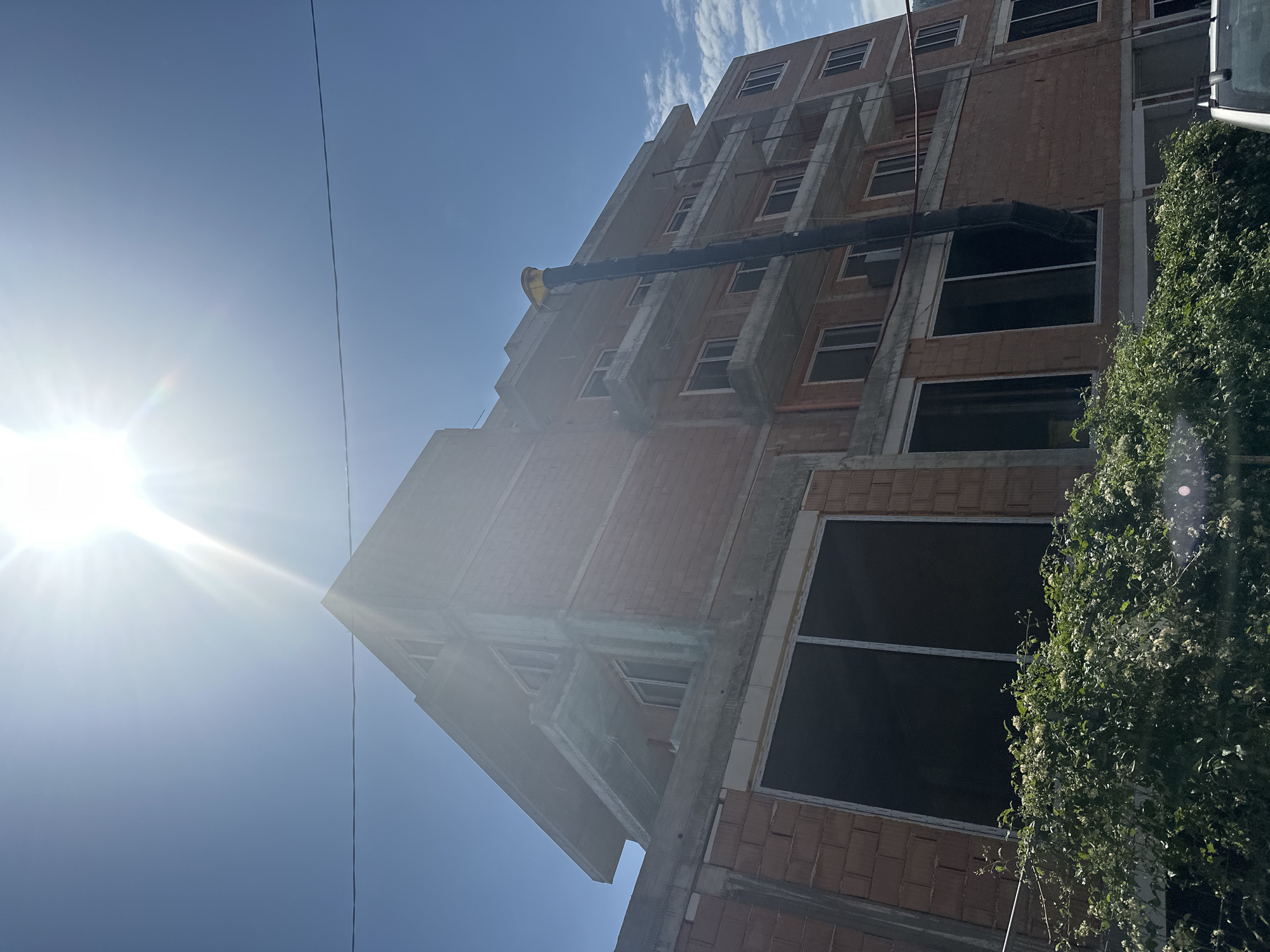 The image shows an unfinished multi-story building under construction on a bright, sunny day. The building, constructed with red bricks and concrete, has several balconies and large dark windows. A tall, black pipe with a yellow device on top extends vertically along the side of the building. In the foreground, lush green foliage with small white flowers obscures the lower part of the structure. A power line is visible in the sky. The scene is set against a clear blue sky with some wispy clouds and a strong sun with visible rays. The location is identified as Vratsa, Bulgaria.
