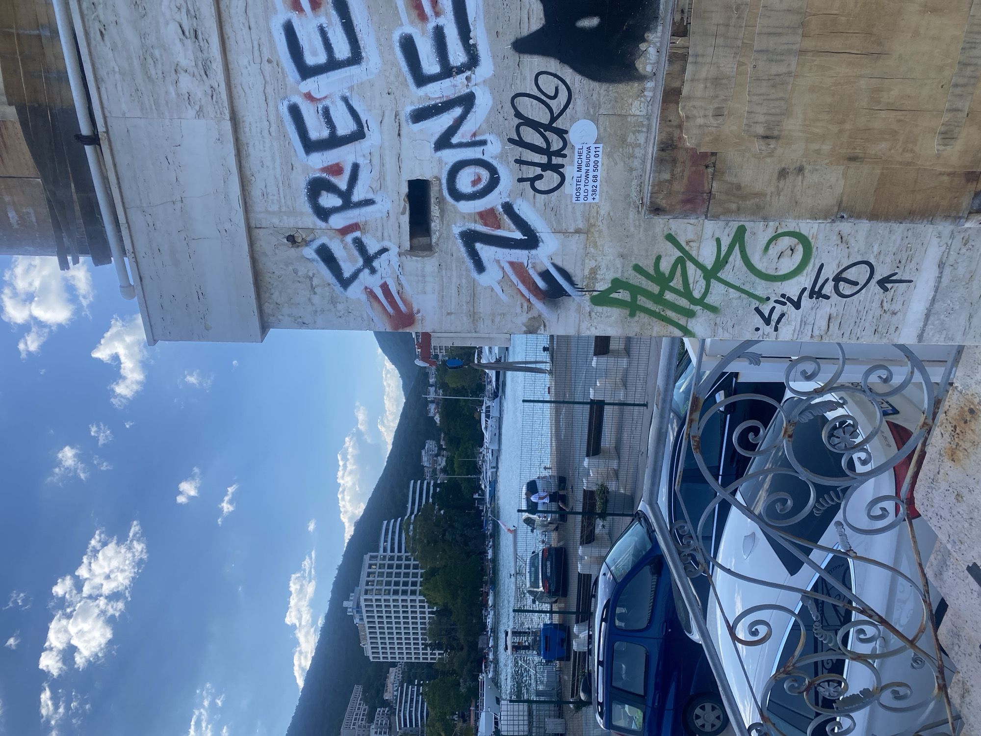 The image captures a view of Budva, Montenegro, likely taken from an elevated position. In the foreground, a stone wall with graffiti, including the words "FREE ZONE", and a metal railing are visible. Beyond the railing, a blue van and a white car are parked. The midground shows a waterfront area with docked boats, a row of parked cars, and buildings along the shore. In the background, a mountain range rises, partially obscured by clouds and buildings. The sky is blue with scattered clouds.