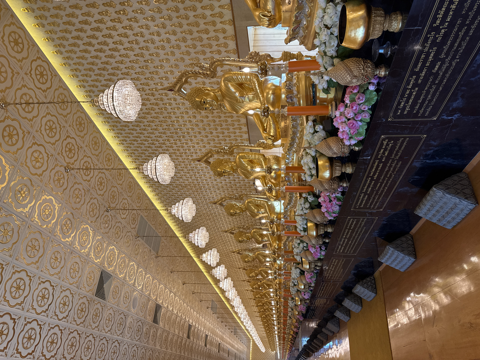 The image shows a hall with a long row of golden Buddha statues. The statues are arranged on a platform with flower arrangements and small woven boxes placed in front. The ceiling is decorated with a pattern of golden symbols and illuminated by rows of chandeliers. Marble panels with Thai script are visible behind the statues. The location is Bang Pu Subdistrict Municipality, Thailand.
