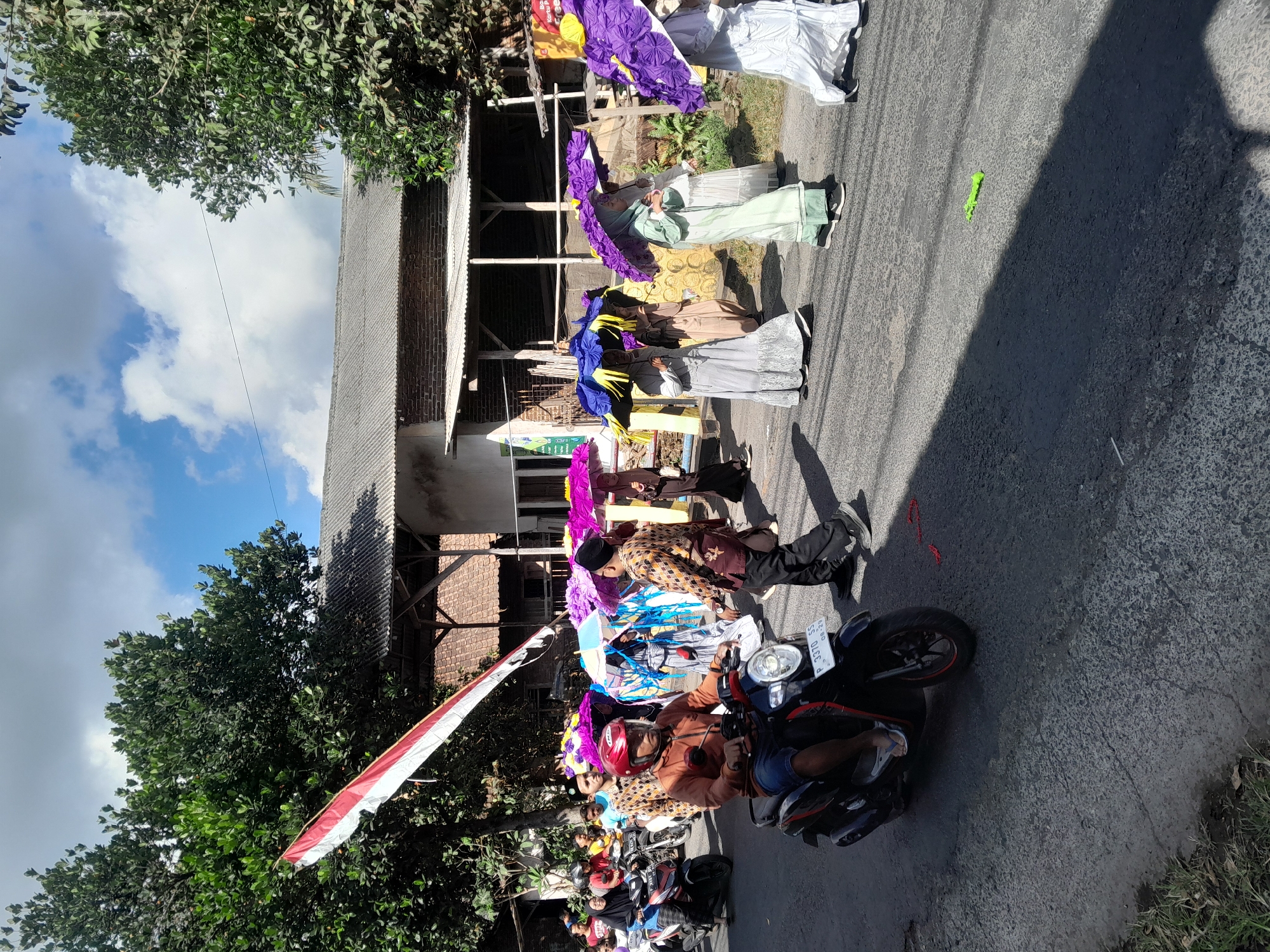 In Gentengwetan, Indonesia, a procession moves along a paved road under a bright, partly cloudy sky. Several people are walking in a line, many carrying large, decorative umbrellas in shades of purple, blue, and yellow. The individuals in the procession are dressed in traditional attire, with some wearing long dresses and headscarves. Behind the walkers, a man wearing a red helmet rides a black scooter, and other motorcycles are visible in the background. Trees line the side of the road, and a building with a corrugated metal roof stands behind them. A red, white, and blue Indonesian flag hangs from a pole on the left side of the image. The overall scene depicts a community event or celebration.
