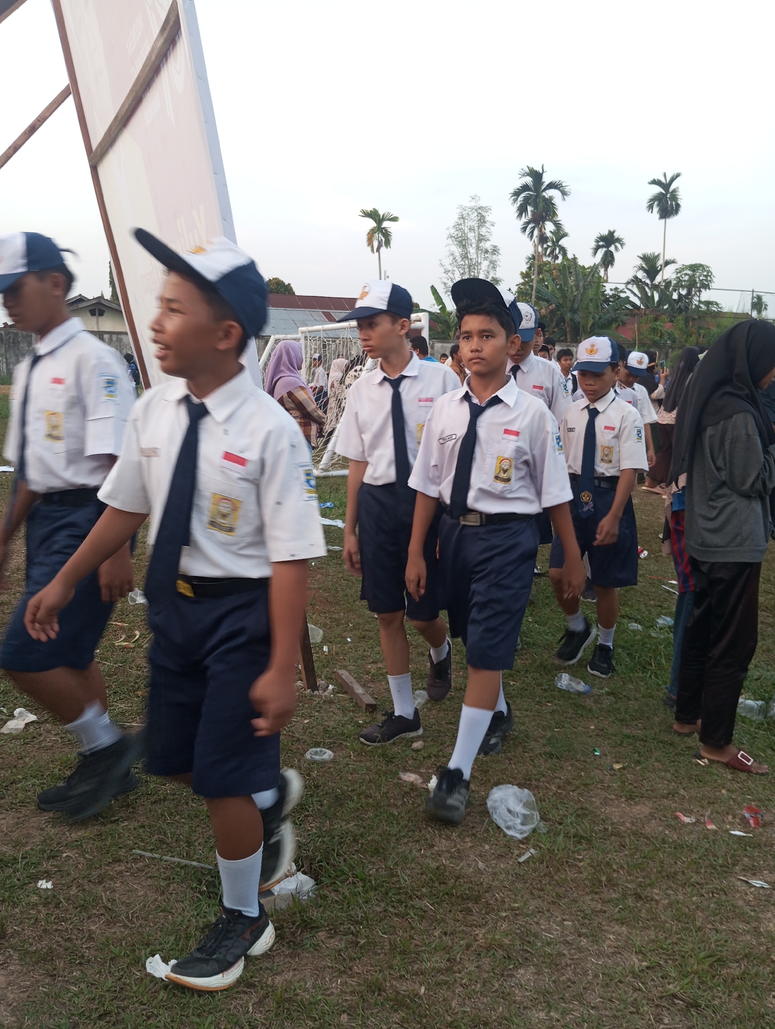 This photograph captures a group of students in school uniforms walking outdoors in what appears to be a grassy field with scattered litter. The students are wearing white short-sleeved shirts, navy blue shorts, and navy blue ties. Many are also wearing navy blue and white caps. The scene suggests a casual outdoor event or gathering. The presence of palm trees and low-rise buildings in the background indicates a tropical or subtropical environment. The image's overall lighting and attire suggest it was taken during daylight hours. While the specific location of Candirejo, Indonesia, cannot be confirmed from visual cues alone, the environment is consistent with a rural or semi-rural setting in Indonesia.