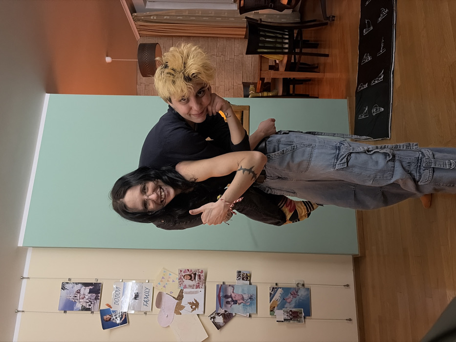 The image depicts an indoor residential scene in Sofia, Bulgaria, featuring two individuals engaged in a playful interaction.
In the foreground, a person with long, dark hair, wearing glasses and a dark top, is carrying another individual on their back in a piggyback ride. This person is smiling broadly at the camera and giving a thumbs-up gesture with their right hand. The second individual, with short, messy blonde hair, a dark top, light-wash baggy cargo jeans, and visible colorful striped socks, is leaning over the carrier's left shoulder, looking towards the camera with a slight smile. A tattoo is visible on the second individual's left forearm.
The room features a light-colored wooden floor. The wall on the left is a light beige and has a vertical string display holding multiple framed photographs and cards, including images of families and text like "DODOVI" and "FAMILY." The central wall is painted a solid mint green. To the right of the central wall, a section of wall with textured, stone-like wallpaper is visible, leading to a dining area. This area has a dark orange ceiling from which a black drum-shade pendant light fixture hangs. A wooden dining table with dark chairs and light cushions is visible in the background, along with a partially visible black mat featuring white patterns on the floor. The scene is illuminated by warm, artificial interior lighting.