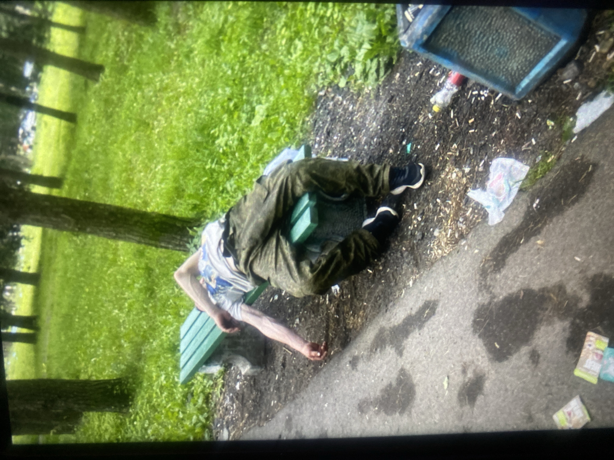 A person is lying face down on a green park bench, with their legs and arms extended. They are wearing camouflage pants and a light-colored t-shirt with a graphic design. Their right arm is hanging off the side of the bench, near the ground. The person's face is not visible. The bench is situated in a park setting with trees and green grass in the background. The ground around the bench is covered in dirt, small stones, and some litter, including plastic bottles and wrappers. A blue trash can is visible to the right of the bench. The overall impression is of someone who has fallen asleep or is incapacitated on the bench. The location is identified as the city of Priladozhsky, Russia.