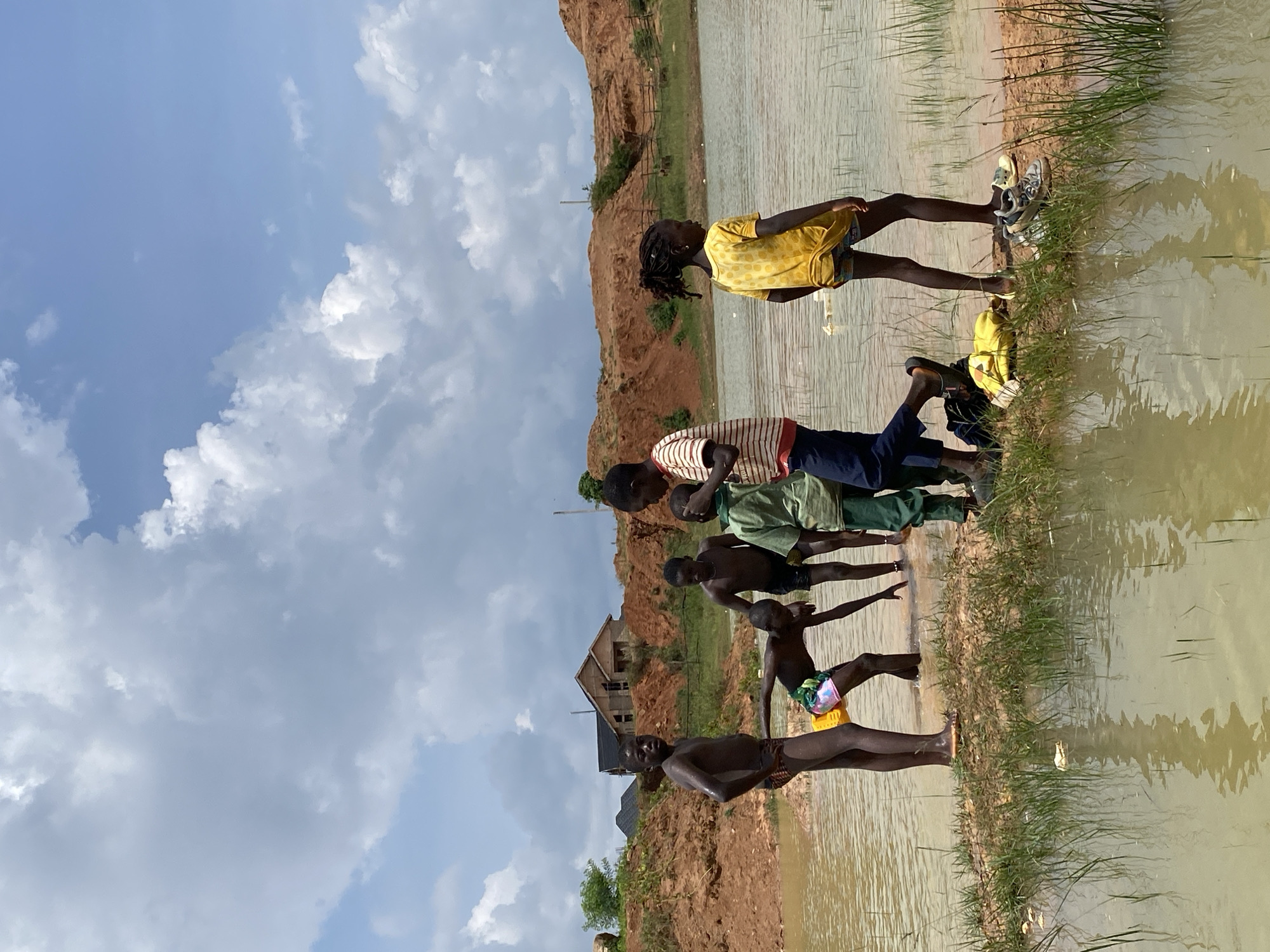 In the city of Bar Jirgi Summa, Nigeria, a group of six children are gathered by a body of muddy water under a partly cloudy sky. The children, all appearing to be under the age of 15, are dressed in casual attire. Two boys in the foreground are shirtless, one wearing patterned shorts and the other wearing striped shorts. Another boy is wearing a green t-shirt and dark pants, and a fourth boy is wearing a red and white striped t-shirt with dark pants. A girl in the background is wearing a yellow t-shirt and patterned shorts. The children appear to be interacting, with some wading in the water. The environment is characterized by reddish-brown earth formations, sparse vegetation, and a large, still body of water. In the background, unfinished buildings suggest ongoing construction. The overall scene depicts a group of children engaged in leisure activity by the water.