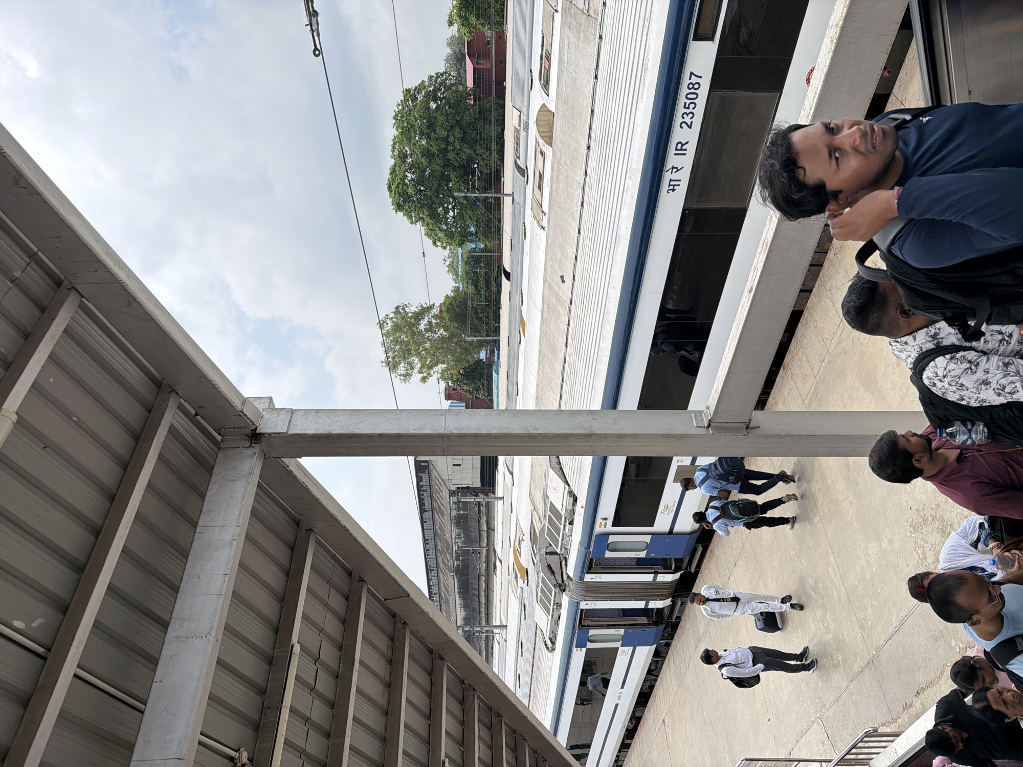 An elevated shot captures a train platform in Lucknow, India, under a partly cloudy sky. A modern blue and white train, identified by the markings "भा रे IR 235078," is parked at the platform. Several individuals are visible on the platform, including people in uniform and passengers with luggage. Some are walking, while others appear to be waiting. A covered walkway with a metal roof extends from the foreground, obscuring part of the sky. In the background, buildings and lush green trees are present. The overall scene depicts a busy railway station environment.