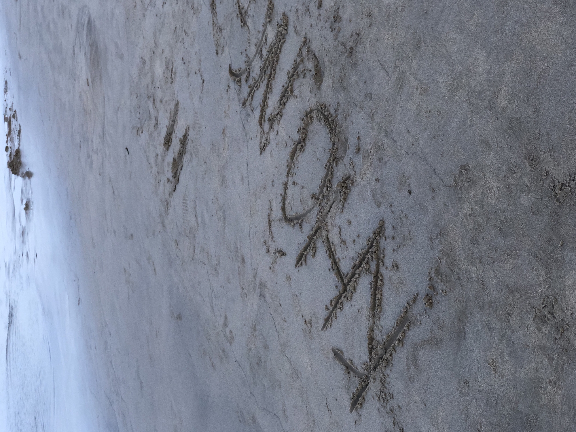 The image shows the word "KHÔNG" written in the sand on a beach. The writing is in large, blocky letters, with each letter formed by a trench dug into the wet sand. The sand appears damp, and there are some small ripples and footprints visible in the background. The ocean is visible in the upper left corner of the image, with gentle waves lapping at the shore. The sky is overcast, casting a soft, diffused light over the scene. The location is a beach in Đà Nẵng, Vietnam.