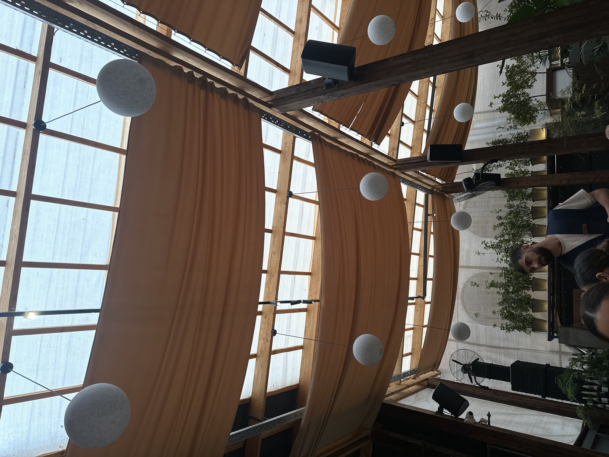 The image shows an interior space with a high ceiling featuring wooden beams, translucent panels, and draping peach-colored fabric. Round, white spherical lights hang from the ceiling. There are black speakers mounted on the beams. In the lower right, a man with a beard and a woman are partially visible. There is also what appears to be a black fan and potted plants visible through the ceiling framework. The location is Yerevan, Armenia.