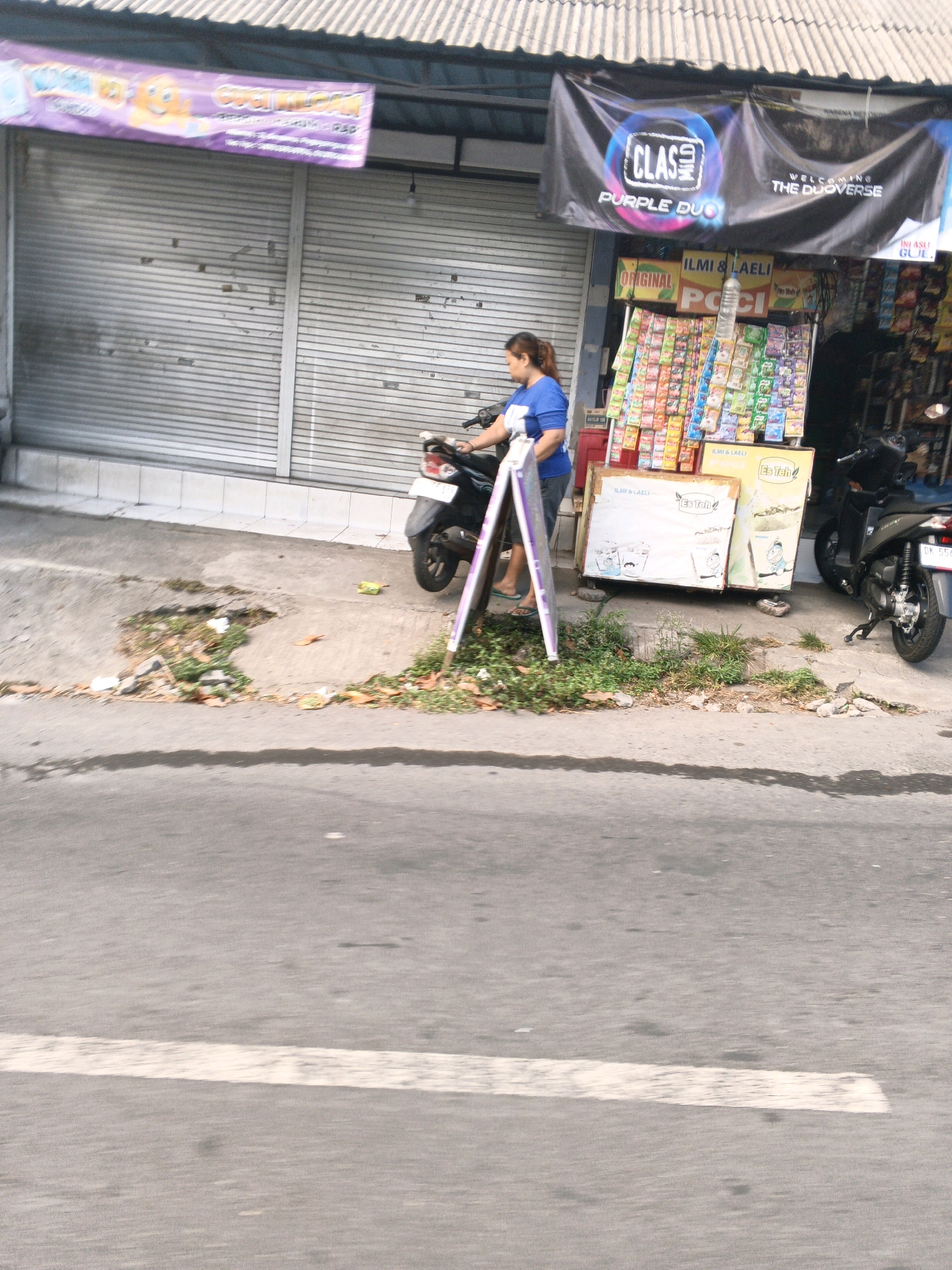 This is a photograph of a street scene in Denpasar, Indonesia. A woman in a blue shirt and dark shorts is standing next to a black motorcycle, facing a store with a roll-up door. In front of the store, a display of packaged goods, possibly snacks or beverages, is arranged on shelves. A white A-frame sign is positioned near the motorcycle. The street in the foreground appears to be asphalt with a white line marking. The background shows the storefront with a corrugated metal roof. There are some plants and debris along the side of the street.
