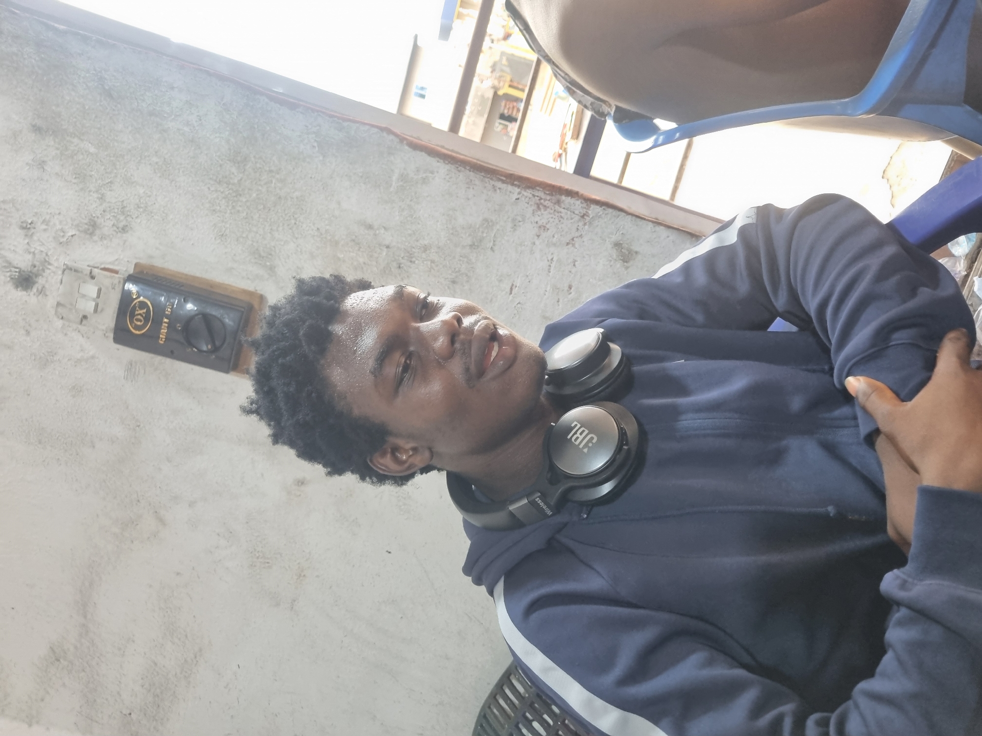 A person wearing a navy blue hoodie with a white stripe on the sleeves and a pair of JBL headphones around their neck sits in a building in the city of Unknown, Unknown. The walls of the building are a textured grey. There is a small electrical box on the wall near the person. There is a hint of other people and a window in the background.