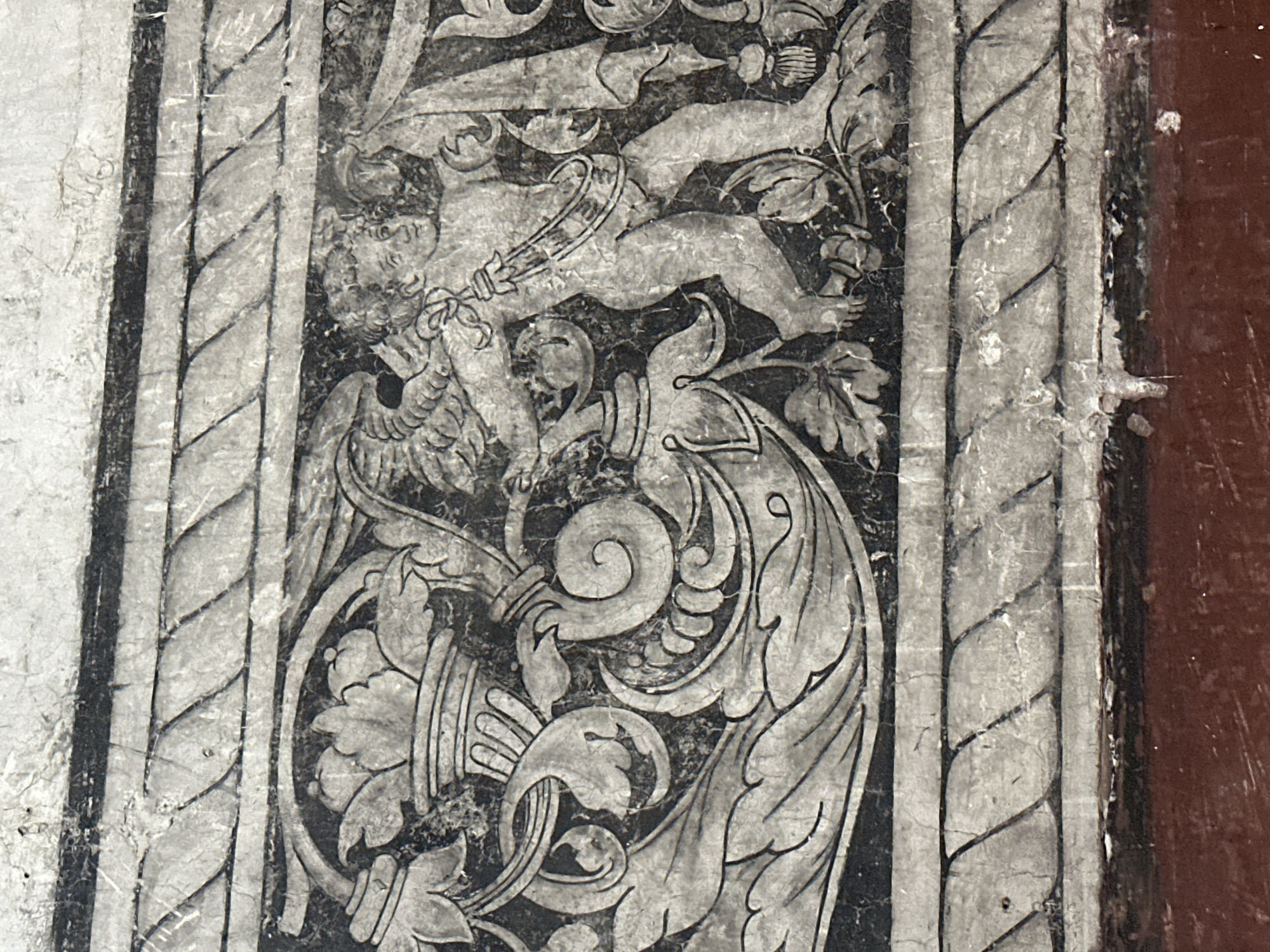 The image shows a fresco detail from the Church of San Miguel Arcángel in Huejotzingo, Mexico. The fresco depicts a cherubic figure, possibly an angel, in a dynamic pose. The figure has wings and is adorned with a sash and a garland. To the left of the figure, there is an ornate urn or vase with floral motifs and scrolling acanthus leaves. The entire scene is framed by a decorative border featuring a rope-like pattern. The fresco is rendered in black and white, with a high level of detail and intricate linework, suggesting a Renaissance or Baroque artistic style. The surface of the fresco shows signs of age, with visible cracks and wear, indicative of its historical significance and exposure to time. The background is dark, providing a strong contrast to the lighter tones of the figures and decorative elements, which are typical of colonial-era church frescoes in Mexico.