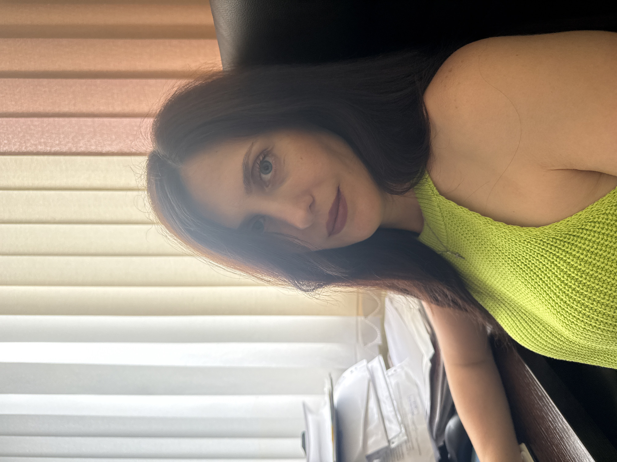 A medium shot captures a dark-haired woman sitting at a desk in an office. She is wearing a lime green knitted top with an open neckline. Her face is turned towards the camera, with her hair falling over her left shoulder. Her eyes are open, and she has makeup on. To her left, a stack of papers and a computer mouse are visible on the desk. Behind her, vertical blinds create a backdrop, with sections of white, beige, and peach colors. The scene appears to be indoors, likely an office environment, possibly located in Kurgan, Russia, though this cannot be definitively confirmed from the image alone.