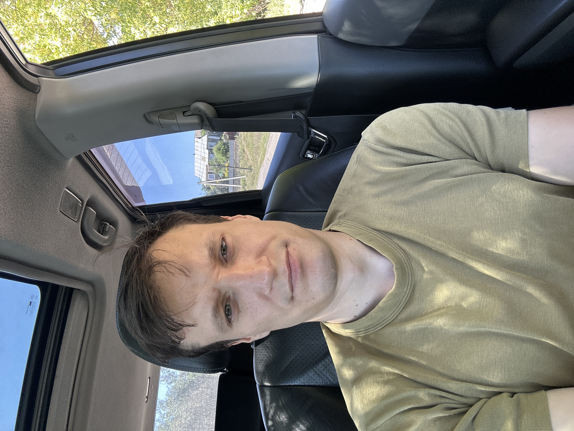 A man with short, dark hair and green eyes is sitting in the driver's seat of a car. He is wearing a light olive green t-shirt. The car has a sunroof open. Outside the car, there is a house with a white roof and a dirt road. Trees and a clear blue sky are visible in the background. The scene appears to be in a rural area.