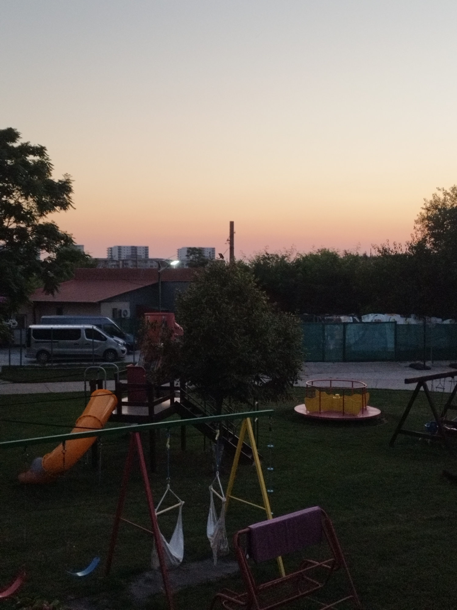 The image depicts an outdoor scene at dusk, likely in a residential area or a small park within Bucharest, Romania. The sky displays a gradient of soft orange and pale yellow hues, indicative of twilight. In the background, several multi-story apartment buildings are visible, suggesting an urban setting.
In the foreground, a playground area is set up with various equipment. There is a bright orange slide, a merry-go-round with yellow seats and a red base, and a swing set with two fabric swings. The ground is covered in green grass.
To the left of the playground, a silver van is parked in front of a building with a red roof. Trees with dark foliage are present throughout the scene, some framing the sky and others lining the perimeter of the playground area. A dark green fence is also visible in the mid-ground. The lighting conditions are low, with shadows beginning to lengthen, contributing to a peaceful, evening atmosphere. No people are present in the image.