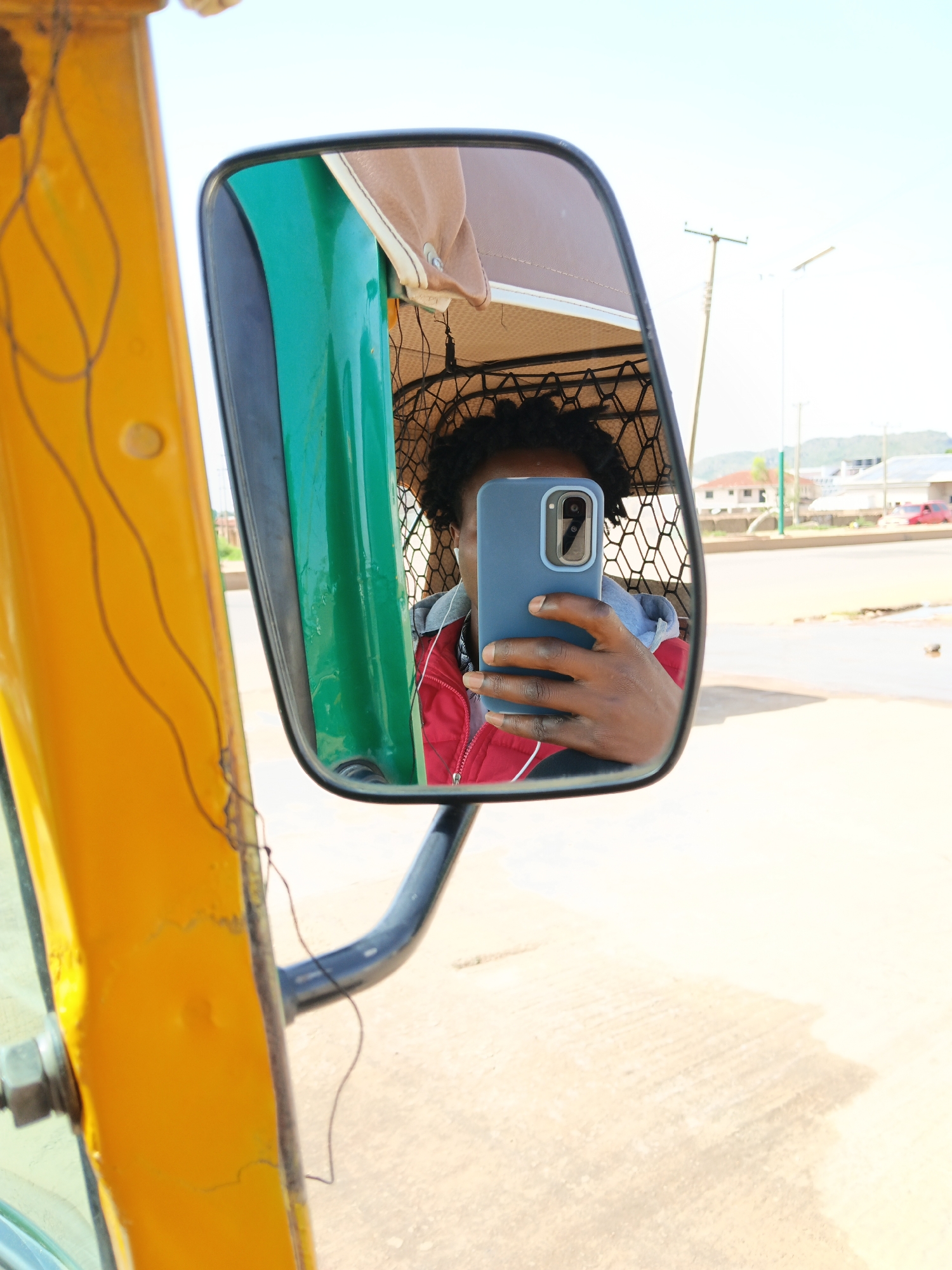 A person is taking a selfie in the side mirror of a yellow vehicle, possibly a tuk-tuk or tricycle. The person is reflected in the mirror, holding a blue smartphone to take the picture. They are wearing a red jacket over a grey hoodie and have curly black hair. The mirror is mounted on a black arm. The background visible in the mirror shows a street scene with buildings, power lines, and a clear sky. The exterior of the vehicle in the foreground is yellow and appears worn. The ground is paved.