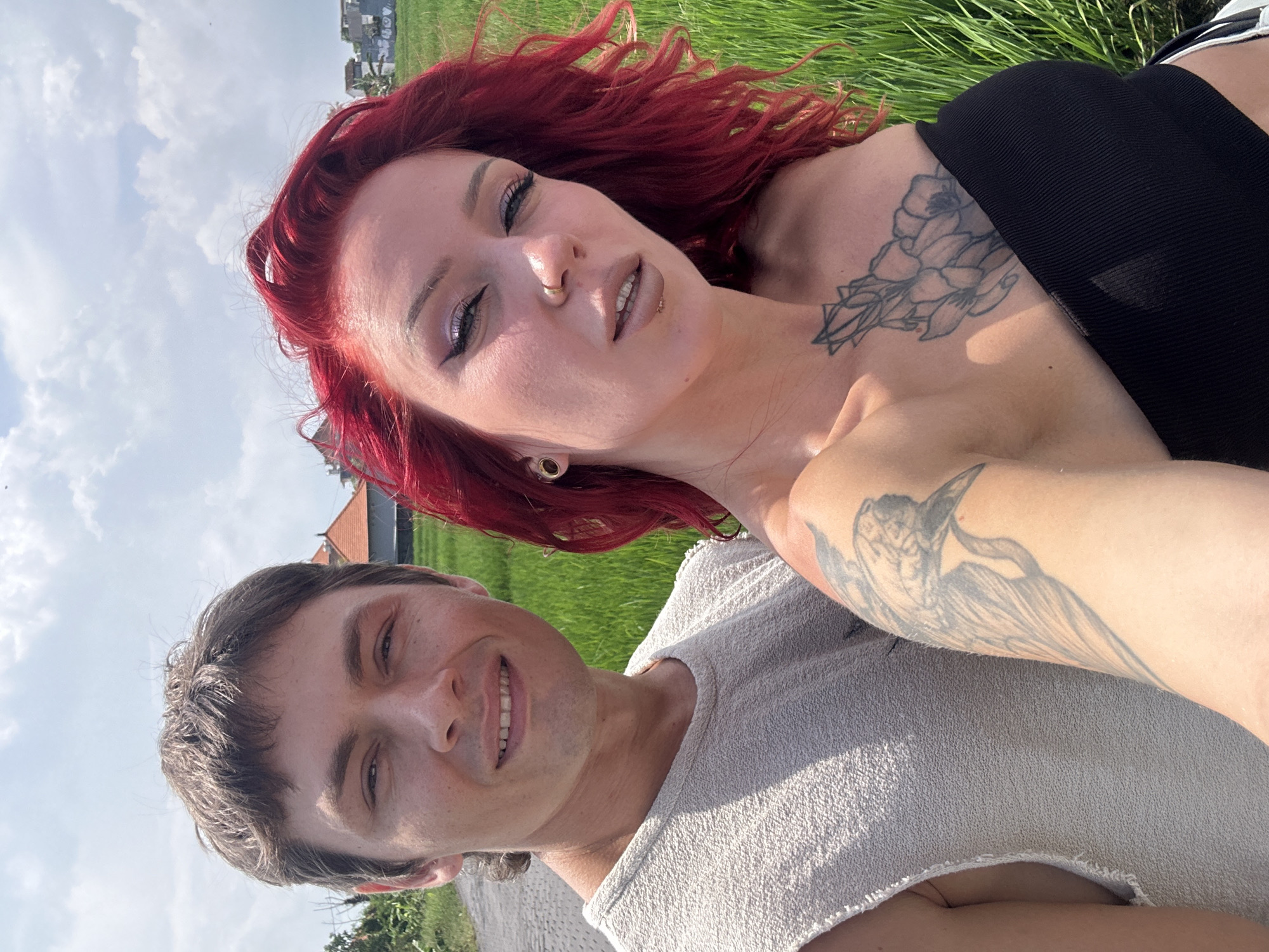 The image shows two people, a woman and a man, lying on a grassy area. The woman has red hair, several facial piercings, and a tattoo on her chest. She is wearing a black top. The man is wearing a light-colored sleeveless shirt and has a tattoo on his arm. The background shows green vegetation, some buildings, and a partly cloudy sky. The location is North Kuta, Indonesia, based on the architecture and the tropical environment.