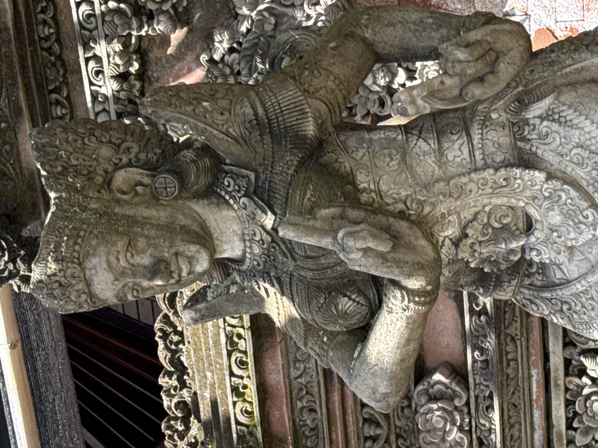 This is a medium shot, eye-level, exterior, natural light photograph of a weathered stone statue of a female figure with elaborate headdress, earrings, and ornate clothing. The statue is positioned in front of a stone wall with intricate carvings. The figure's hands are raised and in a gesture of prayer or blessing, with two fingers extended upward and the thumb touching the first finger. The statue is covered in moss and lichen, indicating age and exposure to the elements. The background shows more stone carvings and architectural details, suggesting a temple or sacred site. The overall impression is one of ancient artistry and cultural significance. The location is in the Ubud District, Indonesia, a region known for its rich cultural heritage and artistic traditions.