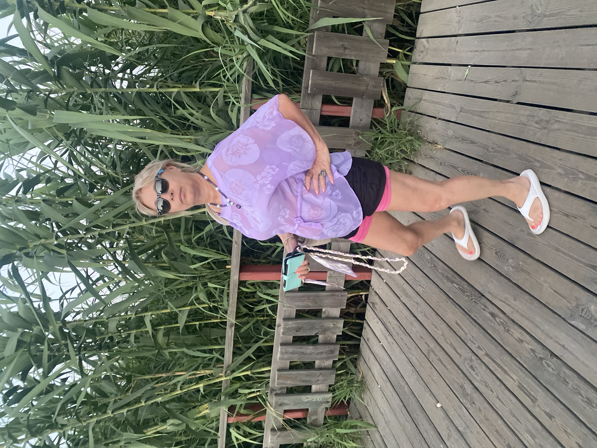 A woman with blonde hair and sunglasses stands on a wooden boardwalk, surrounded by tall, green reeds. She is wearing a sheer, lavender cover-up with a floral pattern over black shorts and pink trim. A beaded necklace hangs around her neck, and she is holding a light blue phone case with a white beaded strap. She has on white flip-flops with red-painted toenails. Her left hand rests on her hip, and her right arm is extended. The scene appears to be outdoors, possibly near a body of water, given the reeds. The lighting suggests it is daytime. The woman's pose and attire suggest a casual, relaxed atmosphere, perhaps a vacation or a stroll along the coast. The boardwalk is made of weathered gray planks. The reeds are dense and fill the left side of the frame, with a wooden fence visible behind the woman. The overall impression is one of a serene, outdoor moment in a natural or semi-natural environment.