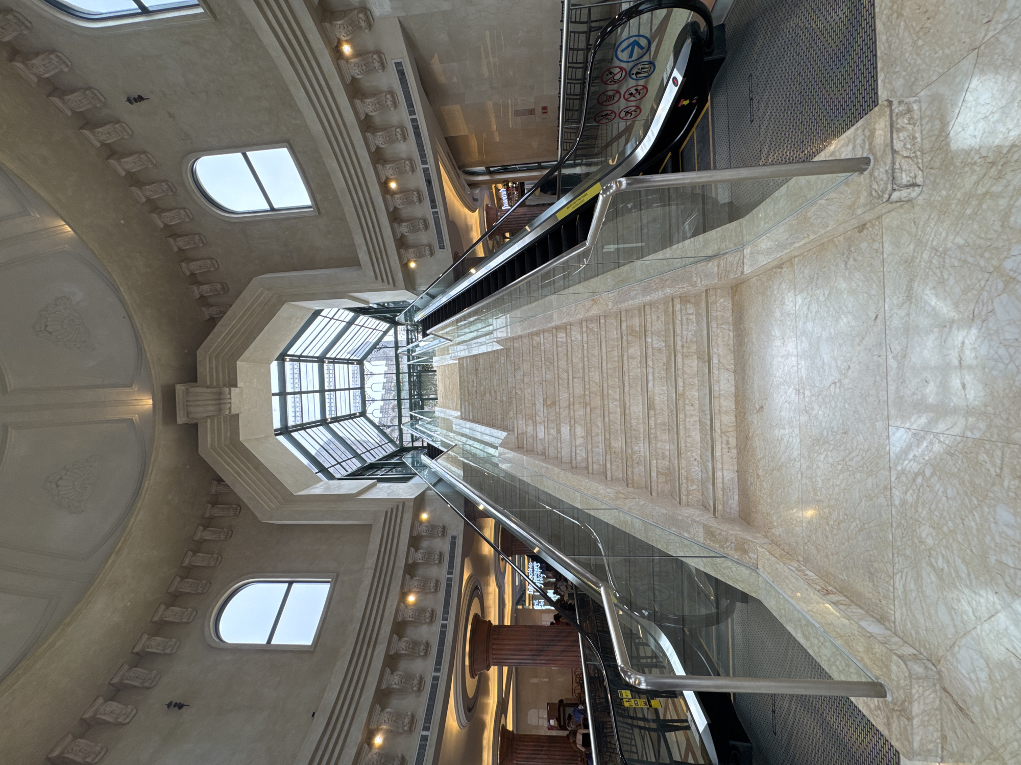 The image depicts the interior of an architectural structure featuring escalators and stairs. The scene is dominated by light beige marble and ornate architectural details. Natural light enters through arched windows on the left and a large, octagonal skylight overhead.
The focus is on the descending staircase and escalators flanked by glass railings. The escalators are operational, with directional signage visible. People are seen on the lower level of the escalator, suggesting the building is open to the public.
The environment exudes a sense of grandeur, emphasized by the high ceilings and detailed embellishments.
Based on the architectural style, material usage, and general layout, the image is most likely taken at Đà Nẵng Railway Station, Vietnam.