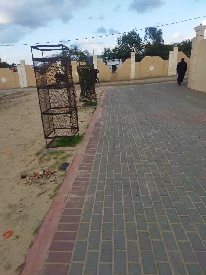 The image captures an outdoor scene, likely a pathway or public area in Khan Yunis, Palestinian Territory, under a partly cloudy sky. In the foreground, a paved path made of grey rectangular interlocking bricks runs diagonally from the bottom right towards the top center. This path is bordered on its left by a strip of reddish-brown bricks, separating it from sandy, uneven ground.
On the sandy ground, to the left of the path, are two large, rusted, rectangular metal wire cages or frames. The one closest to the viewer is empty and appears to lean slightly to the left. Further back, a second similar cage is visible, partly obscured by what appears to be greenery or vines growing within it. Near the base of the first cage, on the sandy ground, is a small pile of debris and trash, including broken pieces of brick and plastic. A bright orange horseshoe-shaped object is also visible on the sand near the bottom left.
In the midground, a person dressed in a long, dark garment (possibly an abaya or thobe) walks away from the camera on the paved path, heading towards the upper right. Another figure in dark clothing is partially visible behind the second wire cage. A low, light brown or beige wall with white decorative accents runs across the midground, acting as a boundary. Behind this wall, trees and bushes are visible. The sky above is light blue with scattered white and grey clouds, and thin utility lines are faintly seen against it. No discernible text is visible in the image.