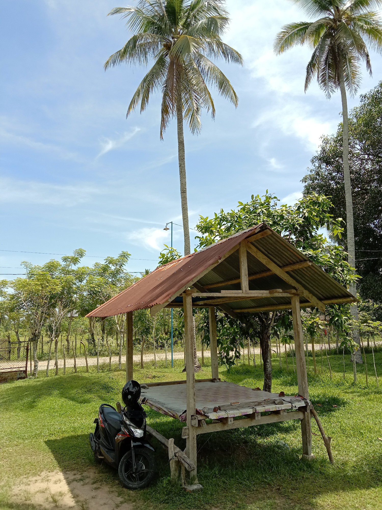 The image depicts a rural scene in Blangkarieng, Indonesia, featuring a small, open-air wooden structure with a rusty corrugated metal roof. A black motorcycle with a helmet resting on the seat is parked beside the structure. Two tall coconut palm trees dominate the background, set against a partly cloudy blue sky. Various smaller trees and shrubs are scattered around the grassy area. A dirt road is visible behind a simple fence.