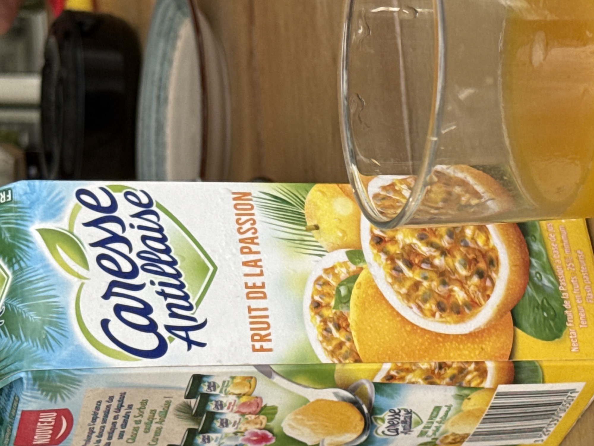 The image displays a carton of "Caresse Antillaise" passion fruit juice, likely situated on a surface in a kitchen or dining area in Saint-Martin, France. The carton's front prominently features the brand name "Caresse Antillaise" in a decorative script, accompanied by "FRUIT DE LA PASSION" indicating its flavor. The packaging is adorned with vibrant illustrations of palm fronds, whole passion fruits, and halved passion fruits revealing their seeds and pulp.
Visible text on the carton further describes the product as a "Nectar Fruit de la Passion à base de jus" (Passion fruit nectar based on juice) with a "Teneur en fruits 25% minimum" (25% minimum fruit content) and notes it is "Flash pasteurisé" (Flash pasteurized). A "NOUVEAU" (New) label is seen on the lower left. Another section of text, partially visible, encourages consumers to "Prolongez l'expérience la douce sensation des fruits exotiques en dégustant notre gamme de Glaces et Sorbets exotiques Caresse Antillaise!" (Extend the experience, the sweet sensation of exotic fruits by tasting our range of exotic Caresse Antillaise ice creams and sorbets!), alongside an image suggesting these other products.
A clear glass, partially filled with a bright orange-yellow liquid, presumably the passion fruit juice, is positioned on top of the carton. In the softly blurred background, a dark bottle and a stack of plates with a distinctive blue rim are visible, hinting at a domestic setting. The lighting suggests an indoor environment, with no specific time of day discernible. No people are present, and the image captures a static arrangement of these items.