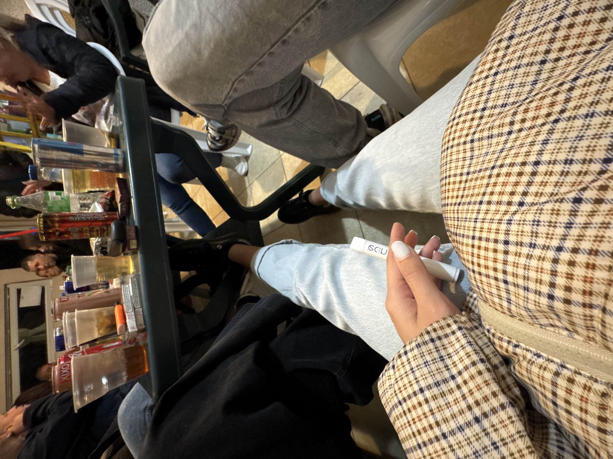 The image captures an indoor scene, seemingly a gathering of people around a table. Several individuals are partially visible; one person wears a black jacket, another grey jeans, and a third a plaid jacket. The table is filled with drinking glasses and bottles, suggesting a social event involving drinks. A white electronic cigarette or vape is held in a hand with manicured nails. The location is identified as Topolovets, Bulgaria, but specific details about the venue are not evident.