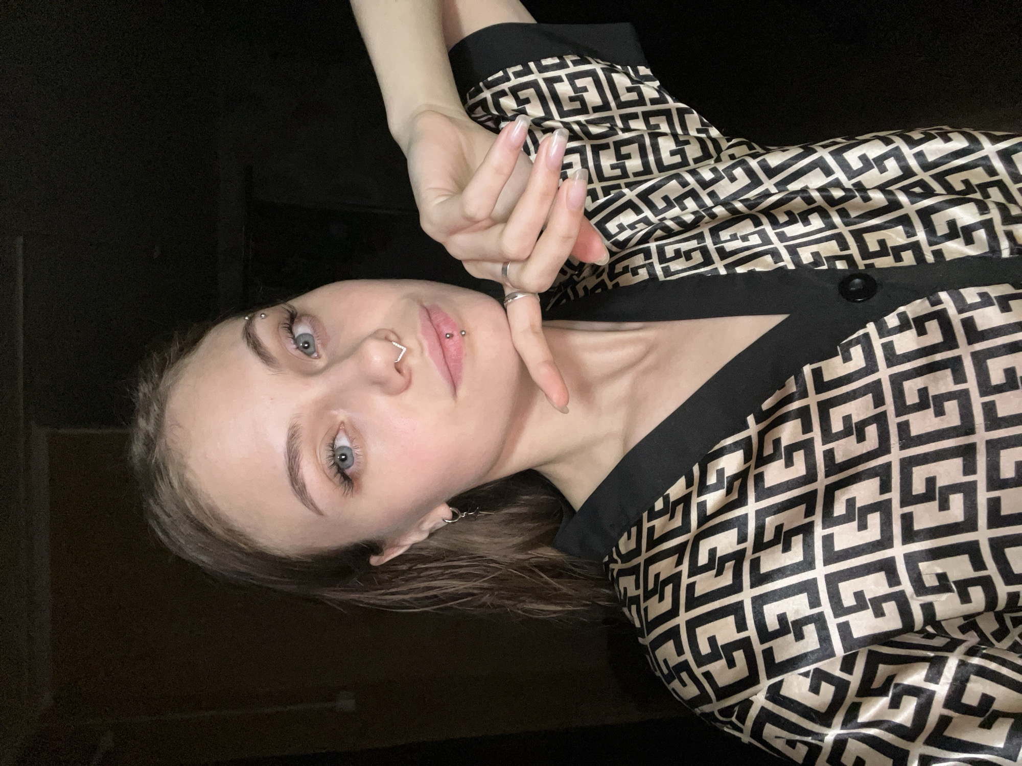 A woman is photographed indoors. She has fair skin, light eyes, a piercing above her eyebrow, a nose ring, and lip piercings. Her long nails are visible. She is wearing a patterned shirt and a ring. The background is dark and indistinct. The location is unknown and cannot be determined from this image.
