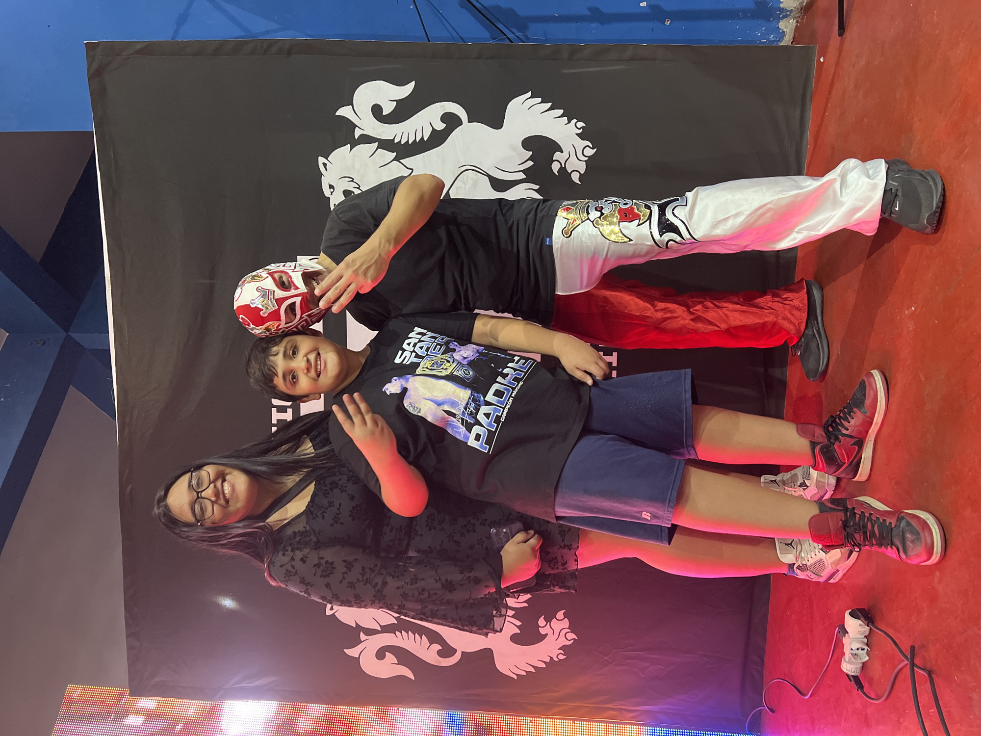 A photo shows three people posing in front of a banner with a white lion symbol. The banner is black, and the location is indoors, likely in Arica, Chile. A woman with glasses is on the left; a boy is in the center, wearing a black t-shirt with an image and blue shorts. A man wearing a red and white wrestling mask and wrestling attire is behind them. The man wears a black shirt with red pants and white wrestling pants. The background includes a blue wall and a red floor. A lighting panel is in the foreground on the left side. A power outlet and plugs are on the floor near the bottom right corner. The overall lighting is indoor with bright, colored lights. The people in the photo are all smiling.