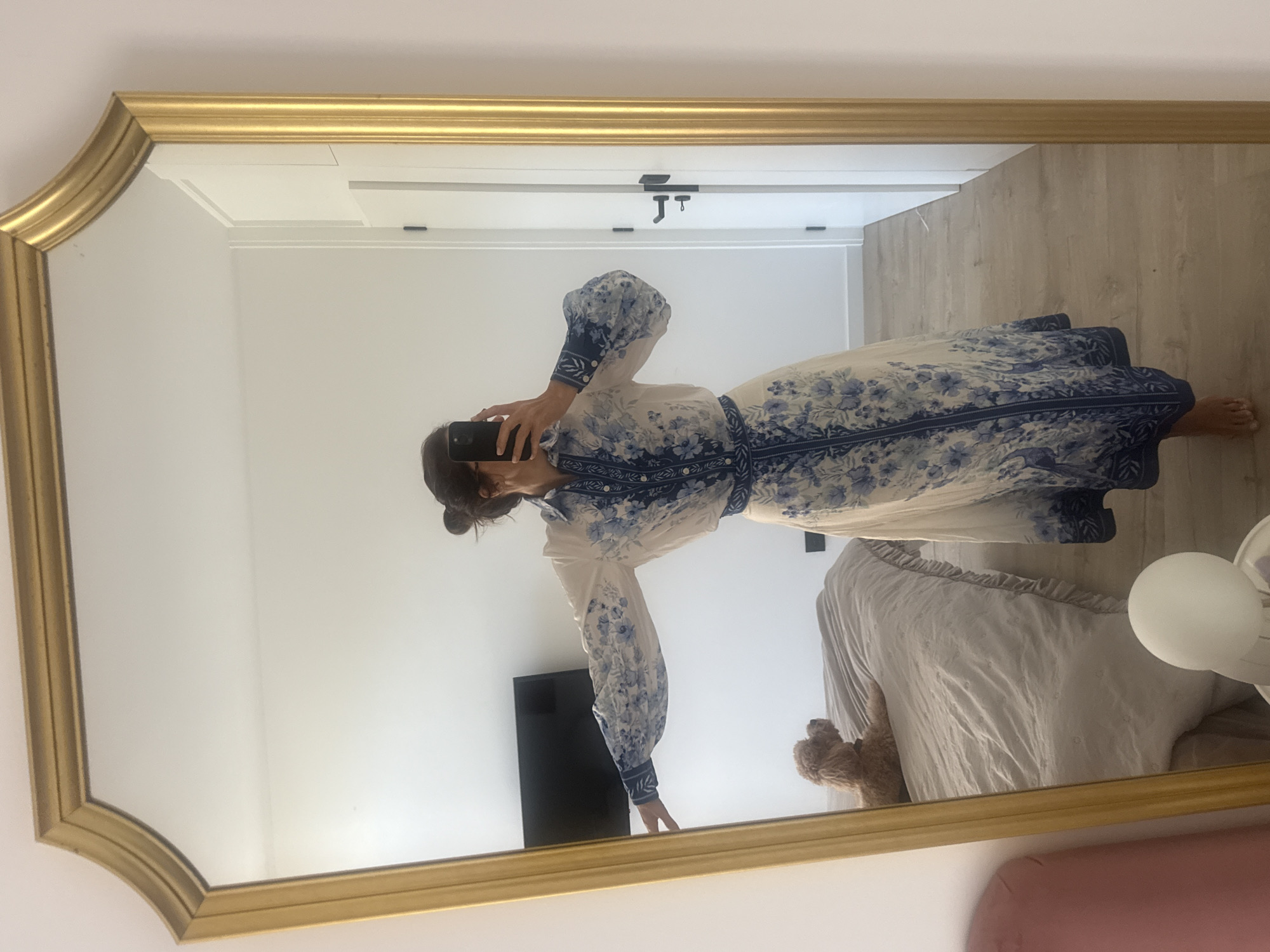 A reflection in a gold-framed mirror shows a woman in a white and blue patterned dress. The woman is holding a phone, presumably taking the picture. The reflection includes a bed with a light-colored comforter, a teddy bear, a nightstand, and a TV. The room has white walls and light wood flooring. A white door with black hardware is visible in the background. The location is not discernible from the image. Vila Nova de Gaia, Portugal cannot be confirmed.