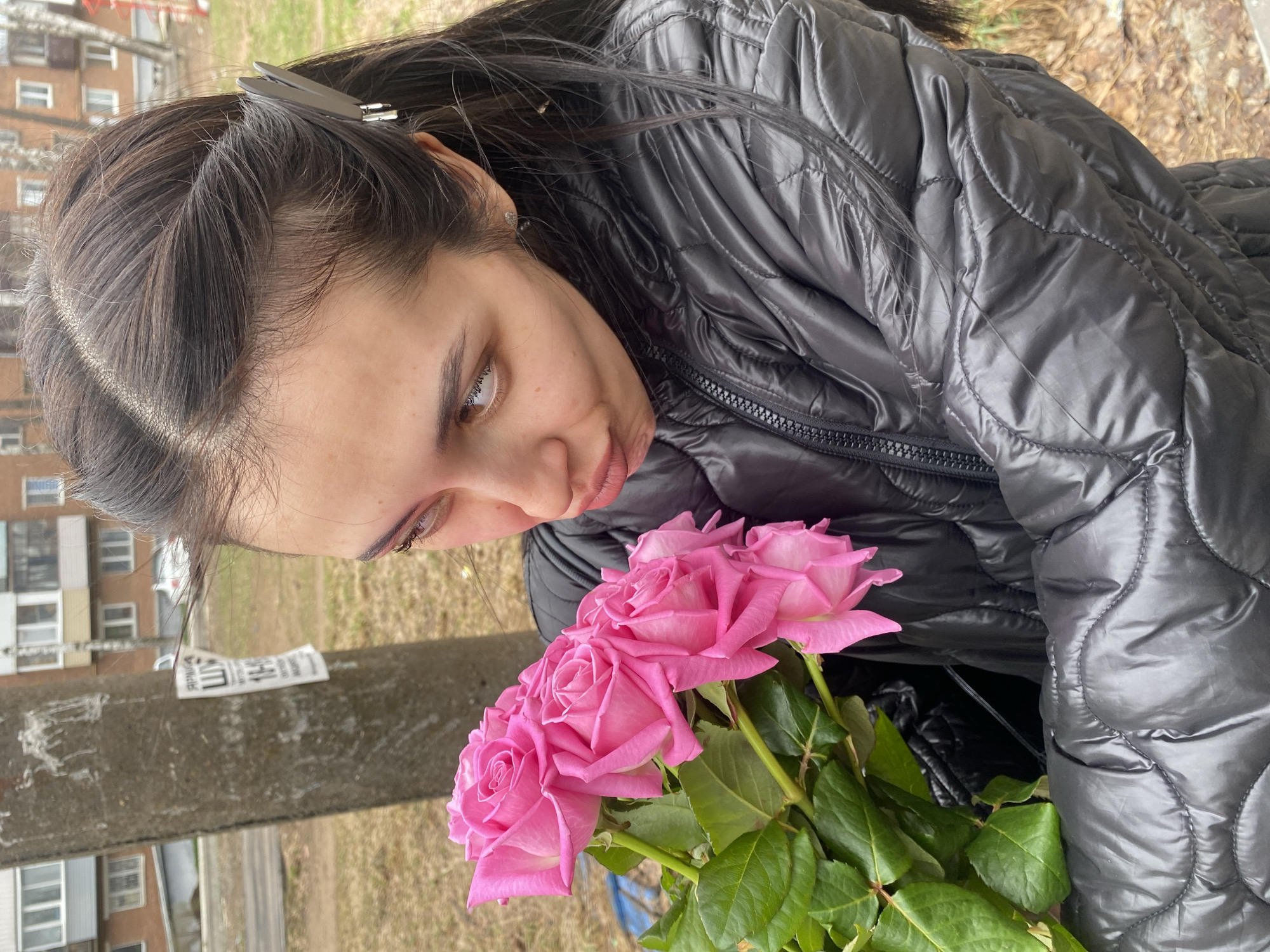 This image, captured in Votkinsk, Russia, features a young woman outdoors, holding and admiring a bouquet of pink roses. She has dark hair, partially pulled back and secured with a gray hair clip, and is wearing a black quilted puffer jacket, suggesting cool weather. Her eyes are light brown, and she has a gentle, slightly pensive expression as she looks down at the flowers.
The scene is set against an urban or suburban backdrop during what appears to be daytime, possibly overcast, given the soft lighting. In the background, a large, multi-story residential building with numerous windows is visible. A concrete pole or beam runs horizontally across the mid-ground, and attached to it is a white piece of paper with Cyrillic text, which appears to be a notice or advertisement. Partially visible characters include "Ш" and "10Ч". The ground beyond the pole consists of bare earth and sparse vegetation, consistent with a late winter or early spring environment. The focus is on the woman and the vibrant roses, creating a moment of quiet appreciation amidst an ordinary city setting.