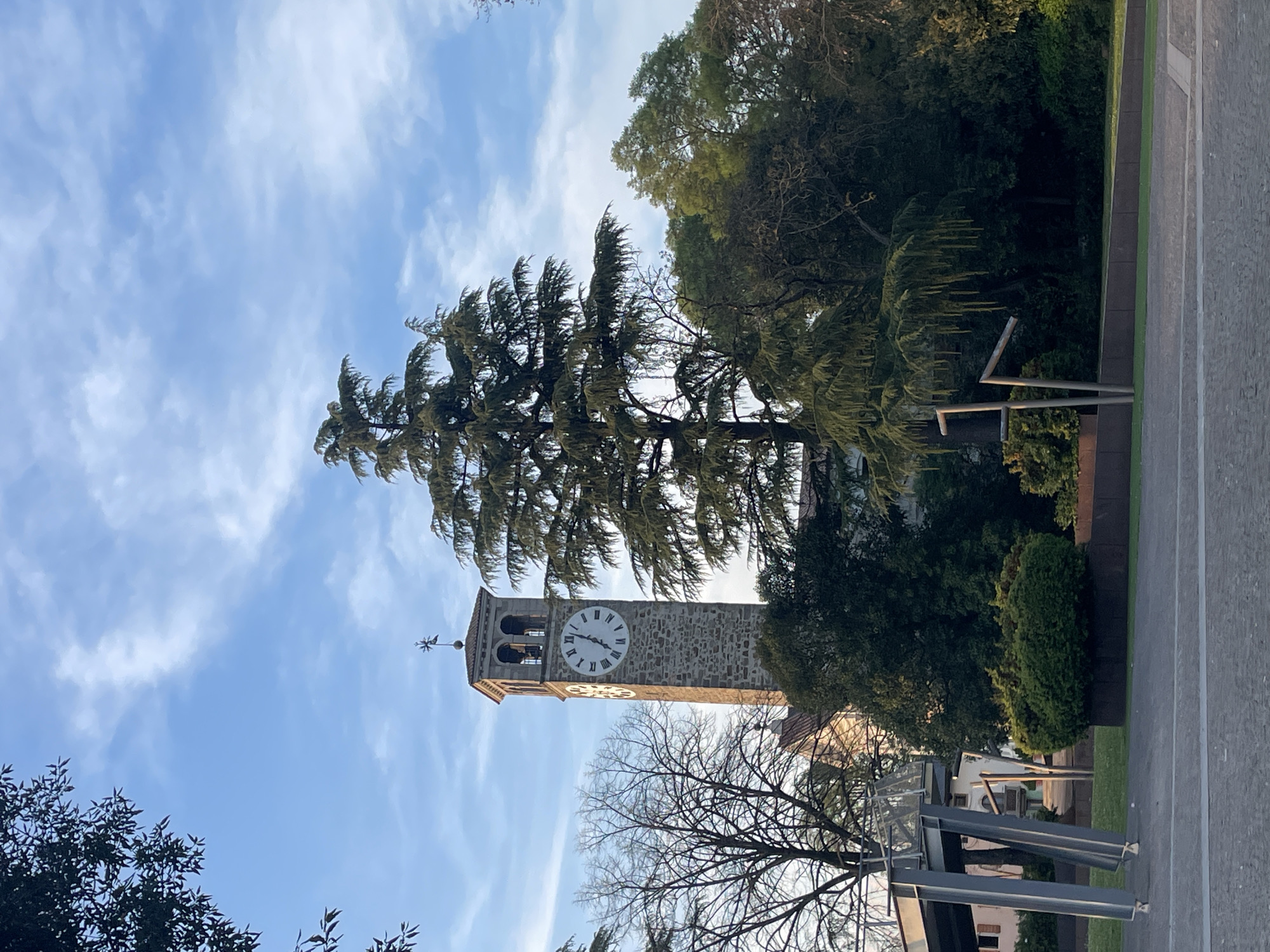 The image captures a view of the bell tower and clock of a stone building, identified as being located in the city of Case / Scjasis, Italy. The tower is prominently positioned with a large clock face visible. The tower is partially obscured by tall green trees with some of them blocking the view from the right side. In the foreground, a leafless tree and a metallic street fixture are visible. The sky is blue with scattered white clouds. A grey walkway is shown on the right side of the image.
