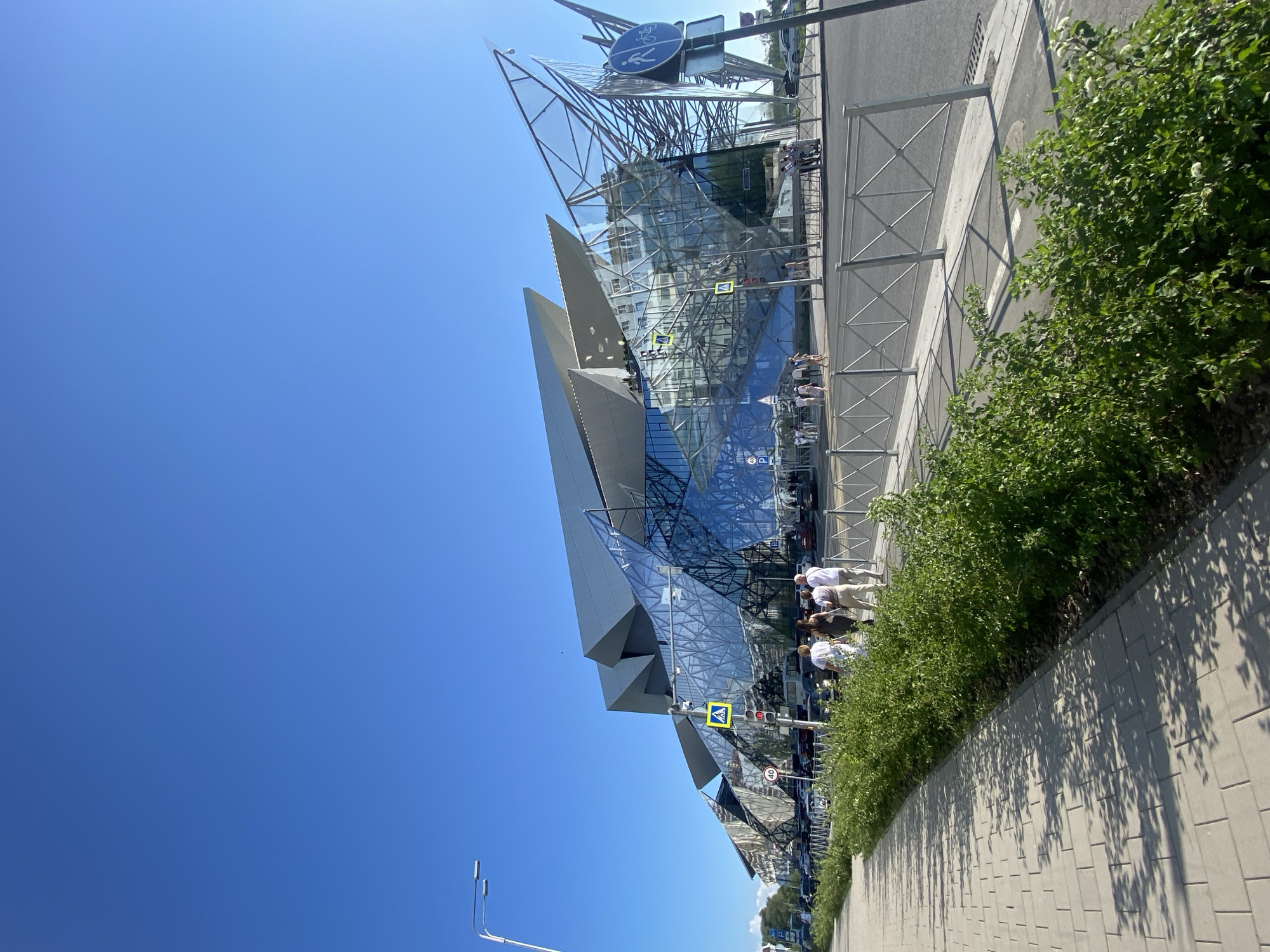 The image shows the Kazan Family Center in Kazan, Russia. The building is constructed of glass and steel and has a modern, geometric design. People are walking outside of the building. The sky is clear and blue. There are bushes and a paved walkway in the foreground.