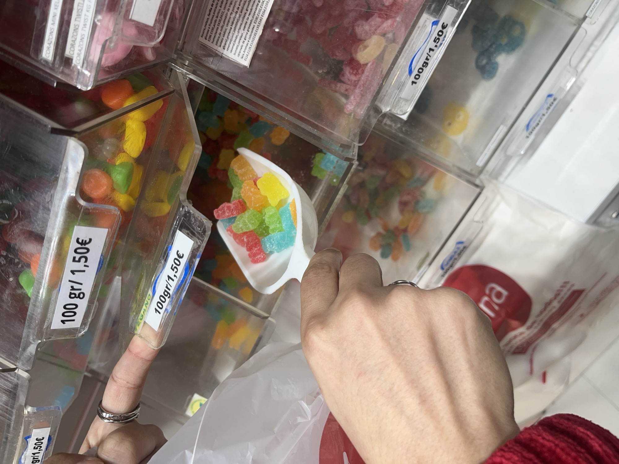 The image shows a person scooping gummy candies into a bag, likely at a bulk candy store. The candies are multi-colored and appear to be in individual bins, each marked with the price of 1.50€ per 100 grams. The label "VEGANO" is visible on one of the bins. The bag being filled has a red logo with the letters "na" visible. The bottom right corner shows part of a red garment, possibly a sweater or shirt, worn by the person filling the bag. Based on the text "A Coruña" on one of the bins, this scene is in A Coruña, Spain. The person is holding a white scoop filled with assorted gummy candies, transferring them into a white plastic bag. A finger with two rings is seen extending towards the candy bins.