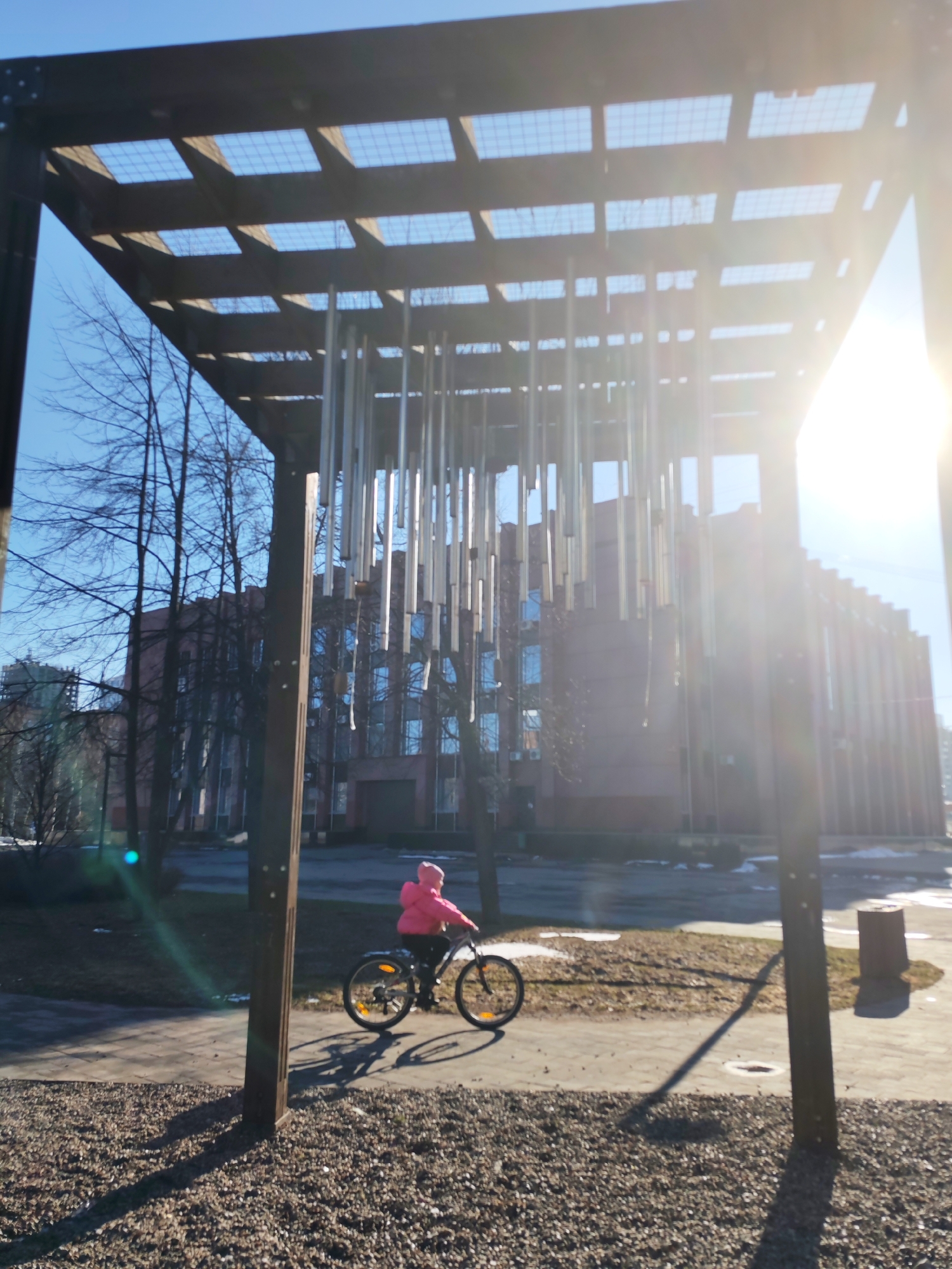 A person in a pink jacket rides a bicycle in a park on a sunny day. The park features a metal structure with hanging metal cylinders, possibly a wind chime. A large brick building is visible in the background. Based on the style of architecture and the presence of the wind chime structure, the location is most likely the "Rodniki" (Springs) park in Izhevsk, Russia. Patches of snow remain on the ground. The ground cover consists of gravel and paved walkways.