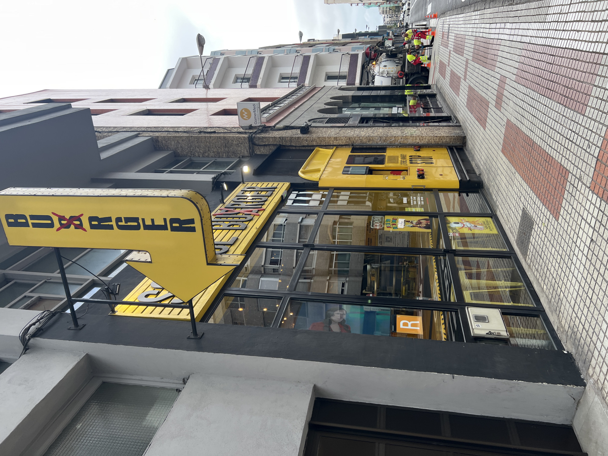 The image captures an urban street scene in Las Palmas, Spain, dominated by commercial buildings under an overcast sky. The view is looking down a street lined with multi-story buildings and a tiled sidewalk.
In the foreground, to the left, a large yellow arrow sign points right, displaying "BURGERS" in black lettering, with a stylized 'X' that appears to have a red cross over it. This sign is affixed to a dark grey building facade featuring large, reflective storefront windows. These windows show reflections of buildings across the street and the sky. Inside the windows, an orange 'R' sign is visible, along with glimpses of interior lighting and a person's reflection. Below the windows, the building's base is light grey.
To the right of the storefront, a yellow food truck or kiosk is parked, bearing the words "STOP" and "FRESH JUICE" on its side. Next to it, several utility or construction workers are visible in high-visibility yellow and red vests, attending to a white and black utility vehicle parked on the street. Orange traffic cones are placed near their work area.
The sidewalk on the right features a distinctive pattern of small white and reddish-brown square tiles. Further down the street, more residential or commercial buildings are visible, including one with a sign reading "GomerMedi." Streetlights extend along the thoroughfare. The overall impression is that of a bustling street with ongoing activity, likely during daylight hours.