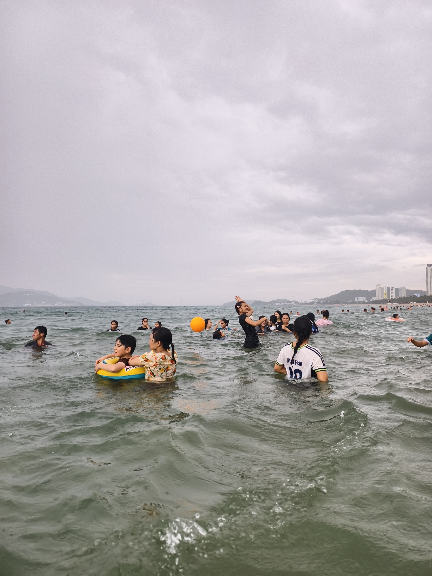 The image depicts a crowded scene of people swimming and wading in a choppy, grey-green ocean under an overcast sky. The sky is a uniform light grey, filling the top third of the frame.
Numerous individuals, appearing to be predominantly Asian, are scattered throughout the water. In the mid-ground, a child is visible in a yellow inflatable swimming ring, which has the text "POOL SCO" partially visible. An adult woman in a floral patterned top is close behind the child, looking towards the left. To the right of the frame, another woman is seen from behind, wearing a white t-shirt with "Mai Trân 00" printed on the back. Further to the left, a man with dark hair is partially submerged, looking towards the right. An orange ball floats among a group of people in the distance on the right side. Many other people are visible further out, enjoying the water, with some appearing to be playing or interacting.
The background shows a distinct coastline. To the left, distant mountains or hills rise from the sea, while to the right, a dense concentration of tall, modern buildings and skyscrapers lines the shore, indicating a city or developed coastal area. The weather appears cloudy or overcast, but the activity suggests a recreational or leisure event at a public beach or resort. The water is visibly turbulent with ripples and small waves.