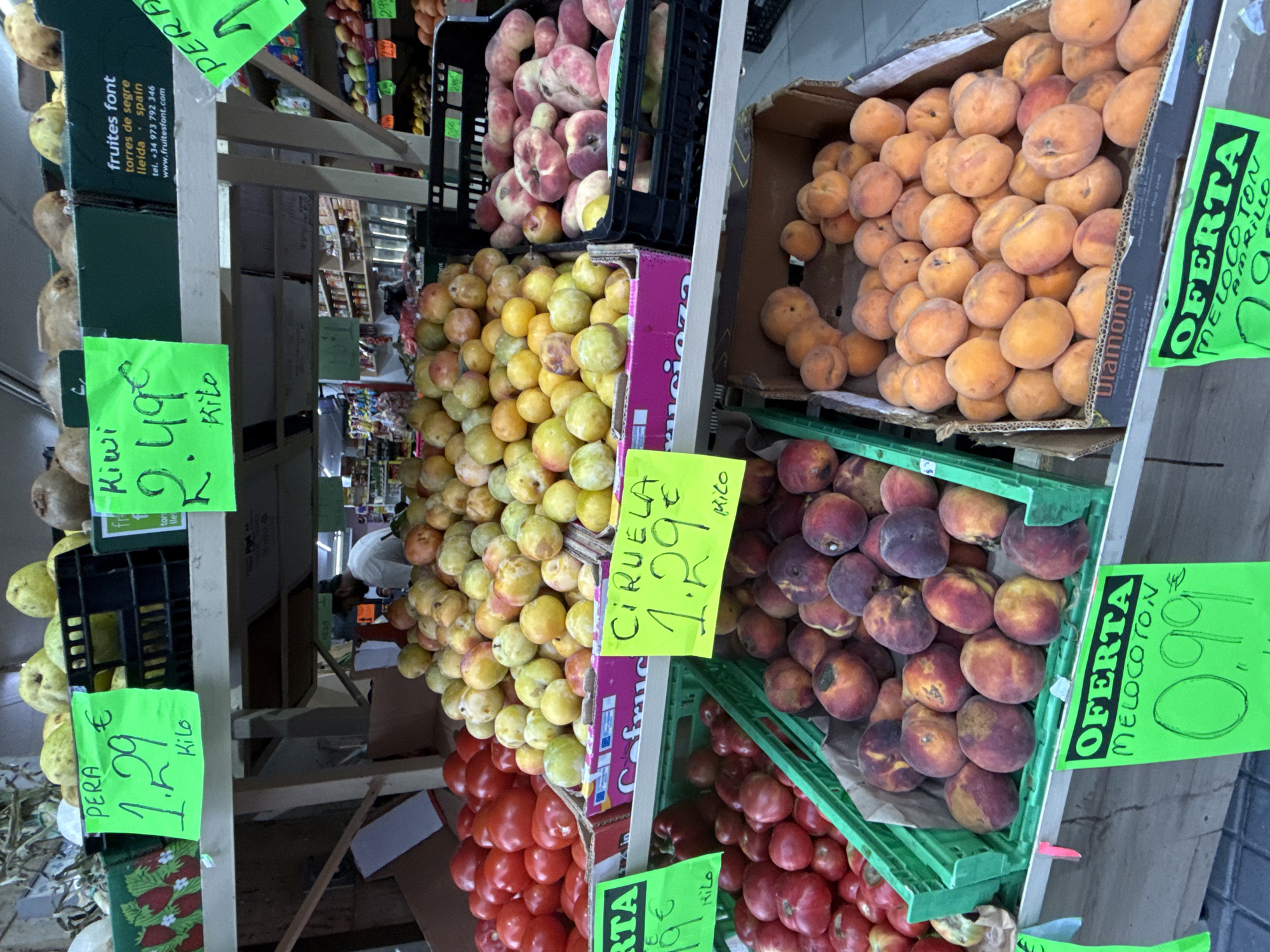 The image displays a produce stand in Blanes, Spain, likely at a local market or grocery store. The stand is stocked with various fruits and vegetables, including tomatoes, plums, peaches, and kiwis. Price tags are visible, indicating prices such as "1.29€" for pears and plums, and "2.49€" for kiwis. A sign also advertises "Oferta Melocoton" for 0.99€. A person, partially visible in the background, appears to be working at the stand, possibly restocking or attending to customers. The overall scene depicts a vibrant and bustling commercial environment where fresh produce is being sold.