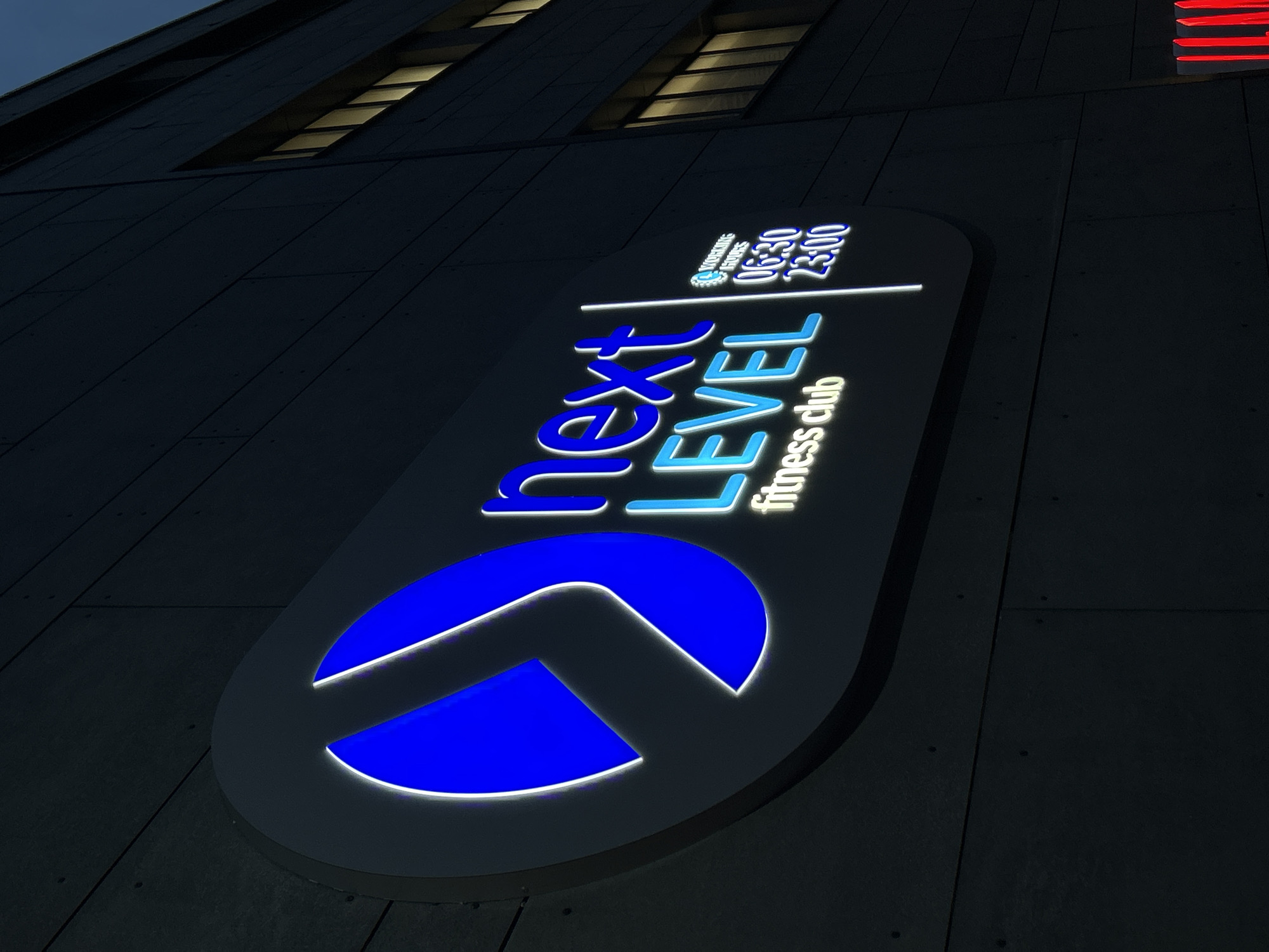 The image depicts a neon sign for "Next Level Fitness Club" in Plovdiv, Bulgaria. The sign is mounted on a building exterior with visible horizontal seams. The sign features a stylized blue logo and the words "Next Level fitness club" in neon blue and white, along with the operating hours "06:30-23:00." There are visible windows towards the top of the building.