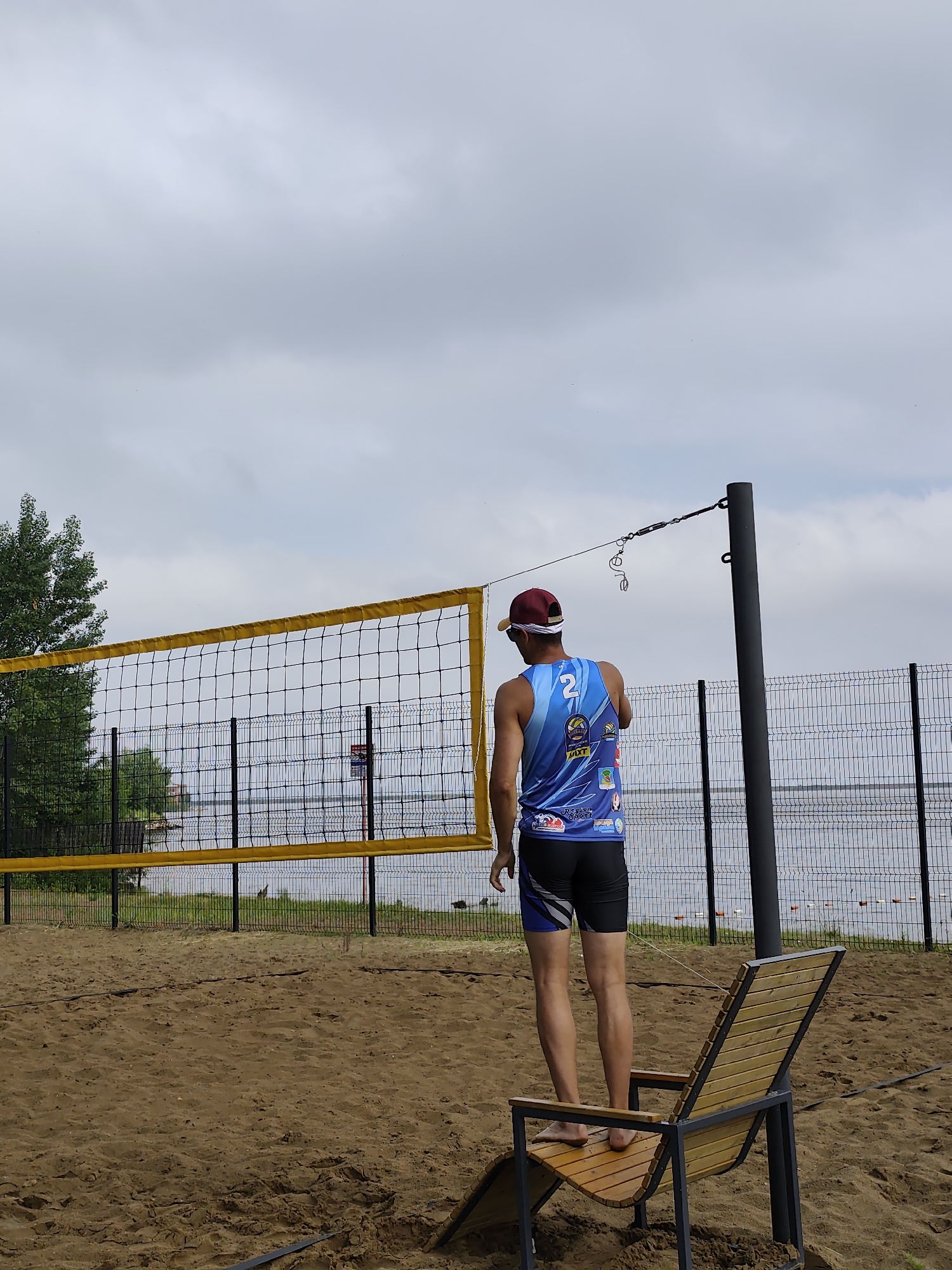 An outdoor, eye-level, full shot captures a person from behind on a sandy beach volleyball court. The individual, wearing a blue and light blue sleeveless shirt with a white number "2" on the back, black shorts with blue accents, and a maroon baseball cap, stands facing a yellow volleyball net. They are barefoot and positioned near a wooden and metal lounge chair on the sand. In the background, a metal fence separates the court from a body of water, likely the Kama River, with trees and some buildings visible on the opposite bank. The sky is overcast, suggesting cloudy weather. This scene is set in the city of Chaikovsky, Russia, specifically at a beach volleyball facility located near the Kama River.