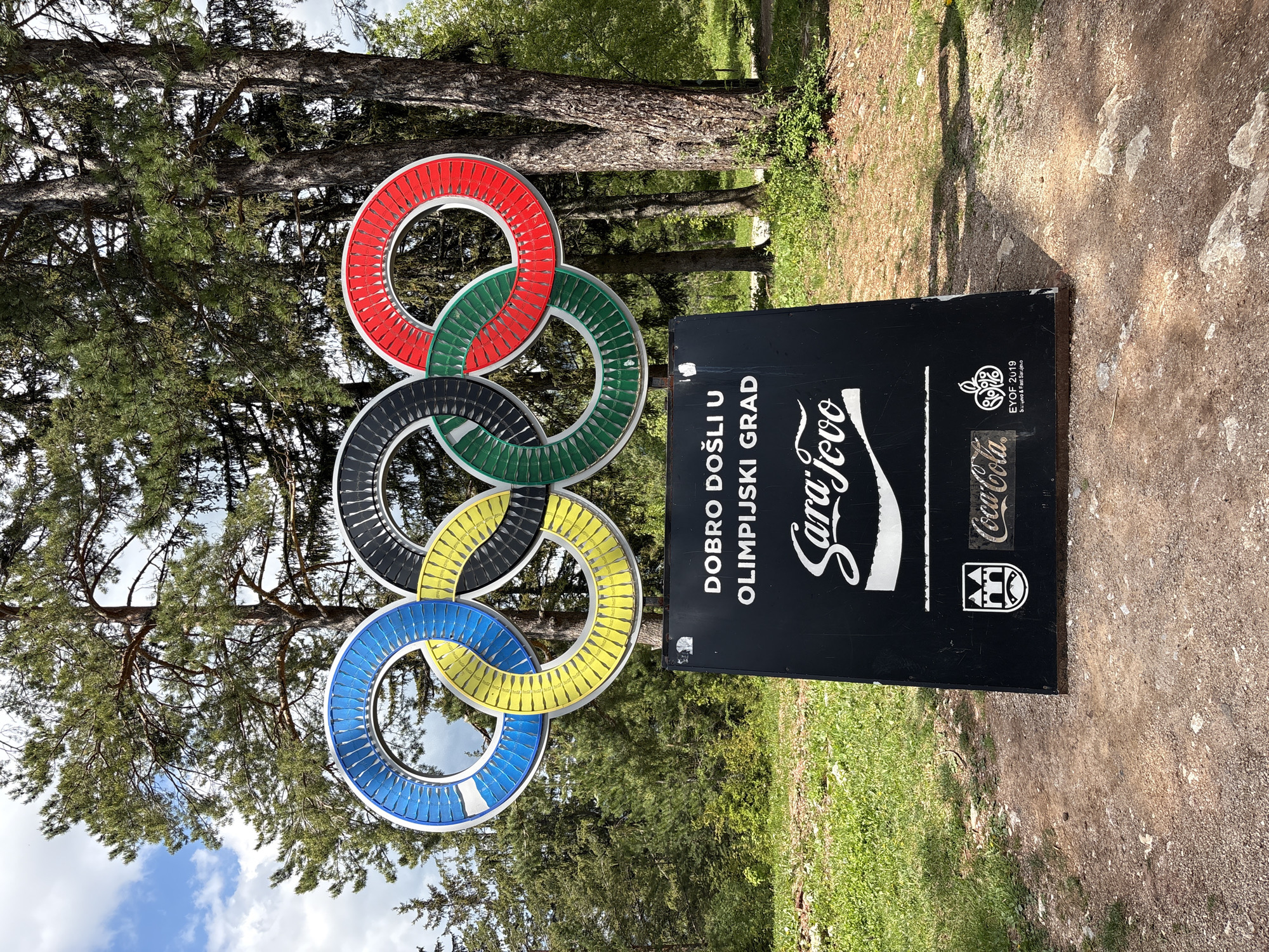 This photograph depicts the Olympic Rings sign in Sarajevo, Bosnia and Herzegovina. The Olympic Rings, colored in red, green, black, yellow, and blue, are situated to the left of a black sign. The sign reads "DOBRO DOŠLI U OLIMPIJSKI GRAD Sarajevo" (Welcome to the Olympic City Sarajevo) and displays the Coca-Cola logo. Below the Coca-Cola logo, there is an emblem for EYOF 2019, which stands for European Youth Olympic Festival 2019. The sign also includes a Sarajevo city logo. The sign is positioned on an unpaved pathway, with trees and greenery in the background. The photograph seems to be taken during the day under natural lighting.