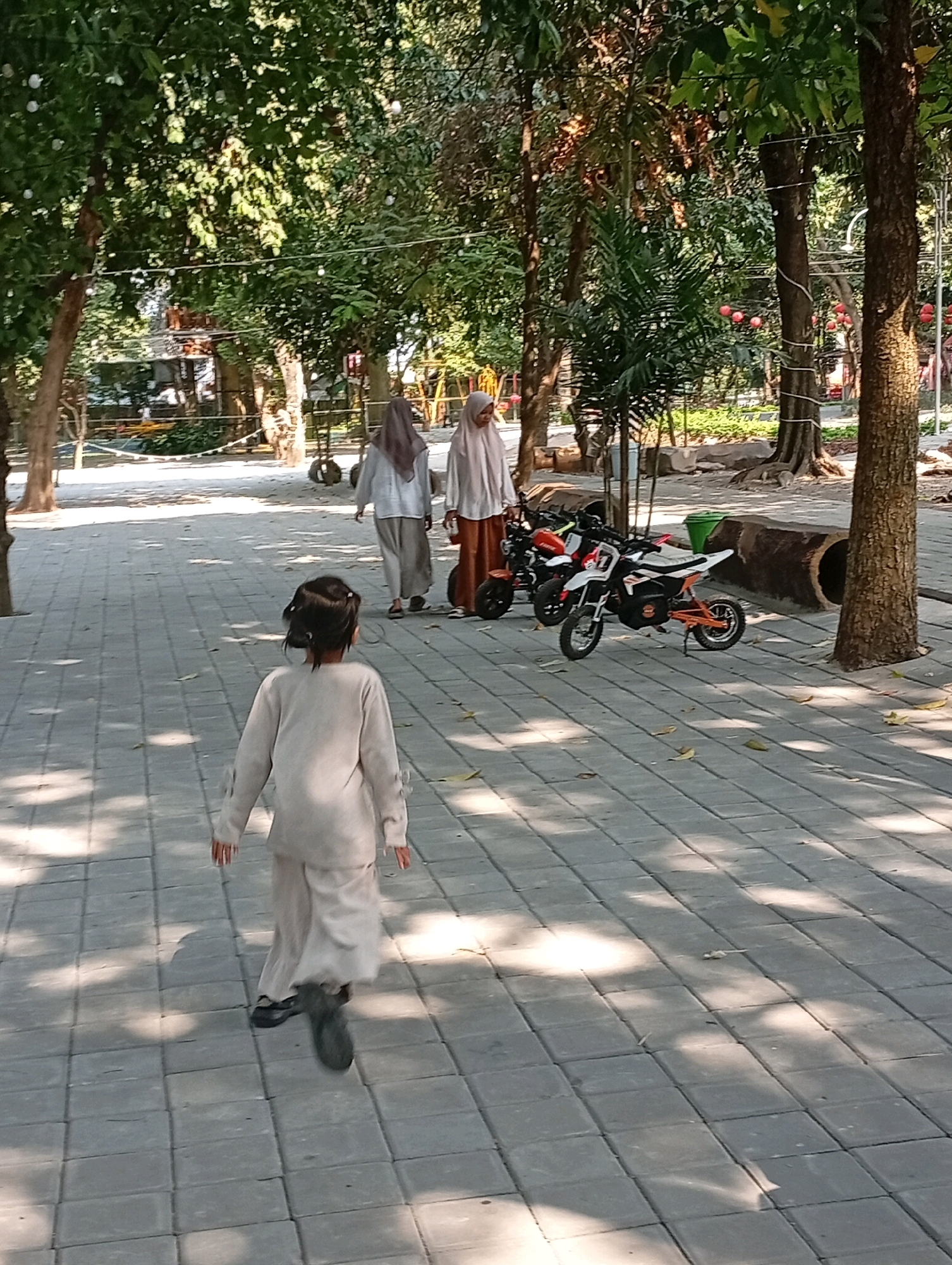 In a park setting in Pandaan, Indonesia, a young girl with dark hair, wearing a light-colored tunic and pants, is walking away from the camera on a paved walkway. She is in the foreground, and her back is to the viewer. Behind her, two women wearing hijabs are walking in the same direction. The woman on the left is wearing a light gray outfit, and the woman on the right is wearing a white hijab and a brown skirt.
Parked on the walkway to the right of the women are two small electric motorcycles. One is predominantly white with orange accents, and the other is orange. There is a green trash bin and a large, hollowed-out log next to the motorcycles.
The scene is bathed in sunlight, casting dappled shadows on the walkway. The trees are lush and green, suggesting it is daytime, likely morning or afternoon, and the weather appears to be pleasant. In the background, there are more trees, some playground equipment, and what appears to be a wooden structure. The overall atmosphere seems peaceful and leisurely. There is no visible text in the image.
