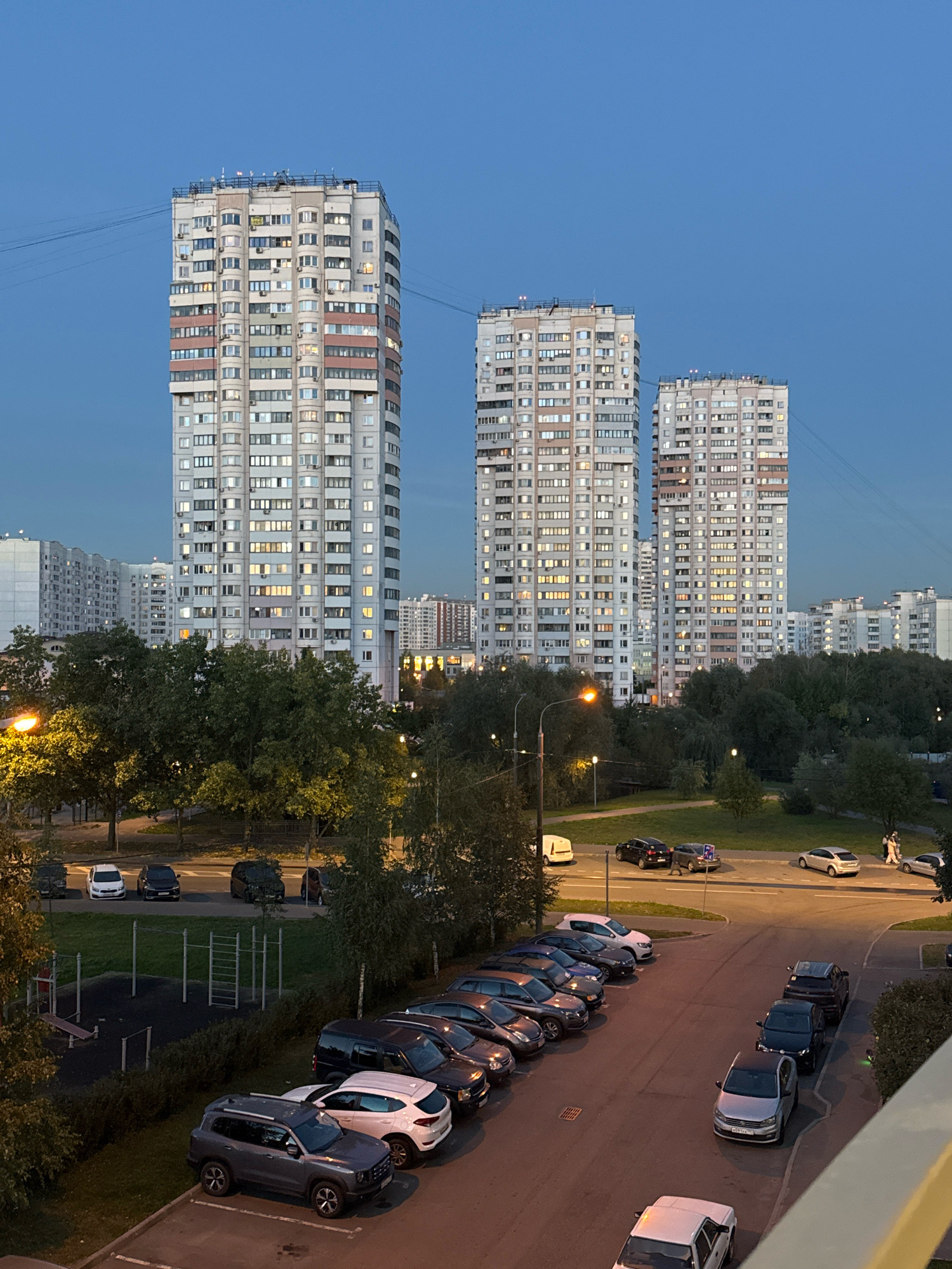 The image depicts a residential area in Zakharino, Russia, at dusk. Three tall, multi-story apartment buildings dominate the background, characterized by their light-colored facades and numerous windows, many of which are illuminated. In the middle ground, a street is visible with several cars parked along its side and others driving. Trees and greenery are interspersed throughout the scene, with streetlights casting a warm glow. In the foreground, a parking lot is filled with a variety of parked cars, and a children's playground with exercise equipment is present. The sky is a deep blue, indicating the transition from day to night. A few people are visible on the street and sidewalk, engaged in walking. The overall atmosphere is that of a typical urban residential environment in the evening.