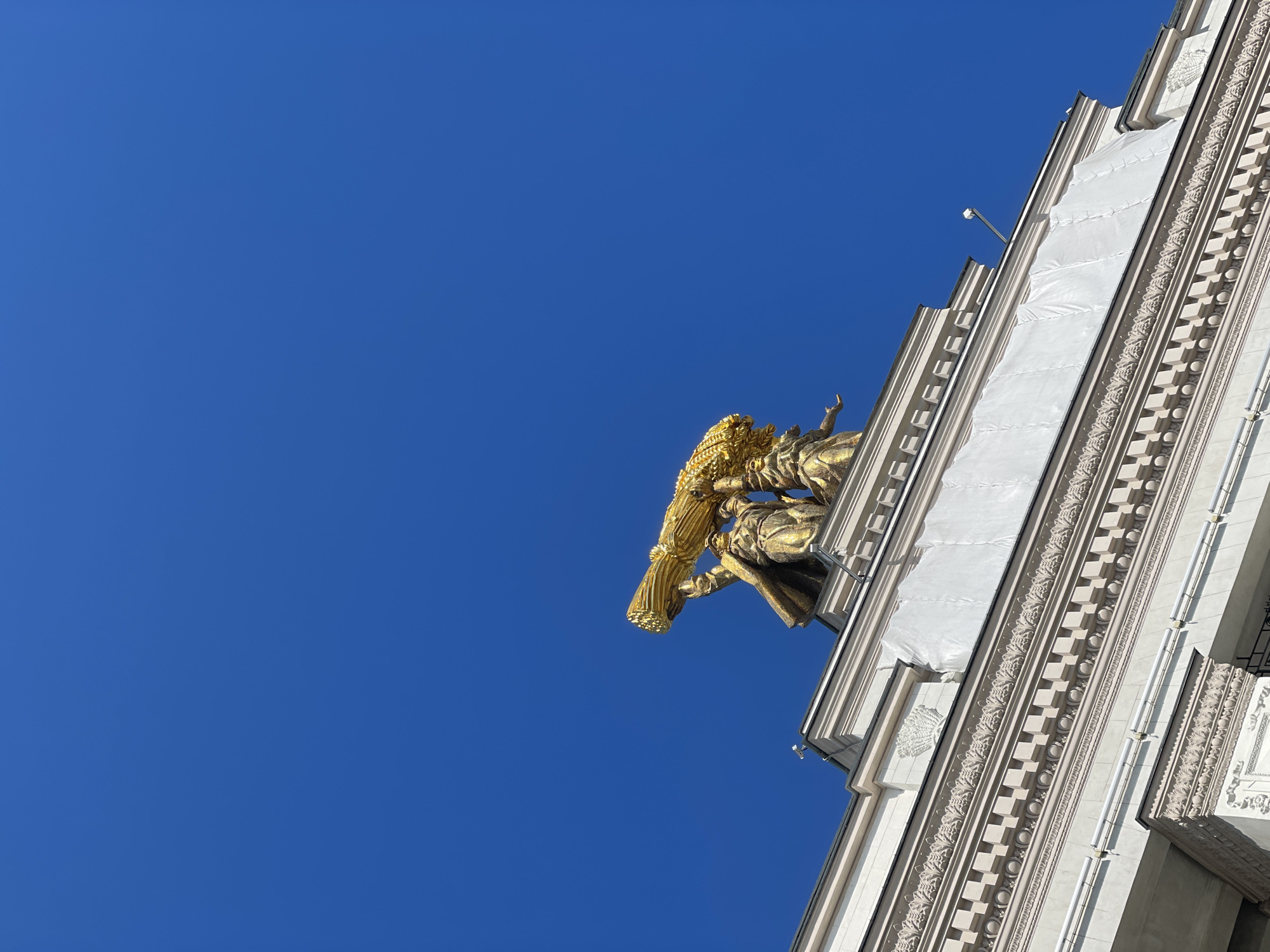 Captured from a low angle, the image showcases a portion of a tall building, likely a landmark in Moscow, Russia, due to its architectural style and ornamentation. The sky, a stark, unblemished blue, dominates much of the frame, providing a backdrop for the building's facade. The building itself is predominantly white, with intricate architectural detailing along its edges, including decorative moldings and patterns. A section of the building is covered with white fabric, perhaps indicative of ongoing construction or restoration work.
The most prominent feature is a gold-colored statue perched on the building. It appears to be a figure holding an object (possibly a sheaf of wheat or a symbolic staff). The statue is highly detailed, showcasing the craftsmanship involved in its creation.
Overall, the image presents a juxtaposition of the architectural grandeur of a Moscow building against the simple backdrop of a clear sky, highlighting both the artistry of the statue and the intricate details of the building's construction.