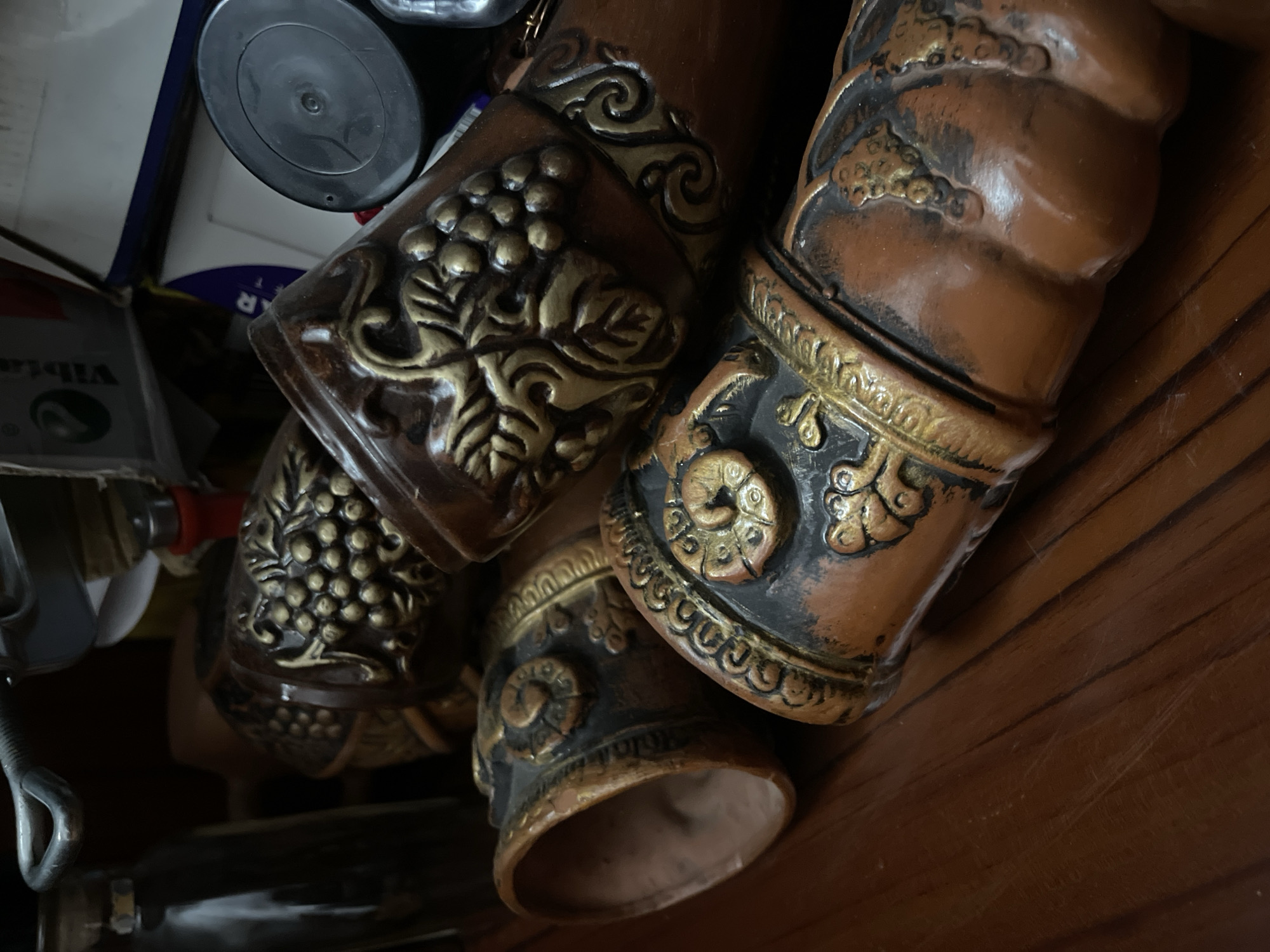 The image shows a close-up of several cylindrical, possibly ceramic, decorative objects with a brownish-orange base color and elaborate gold and black embellishments, including designs that appear to be grapes and leaves. The objects are clustered together on a wooden surface. A mix of other objects, including containers and metal parts, is partially visible in the background, adding to a cluttered impression. Based on the overall imagery and common souvenir items, it is plausible to guess that the image was taken in a souvenir shop. However, without specific details, the location cannot be definitively pinpointed to Tbilisi, Georgia.