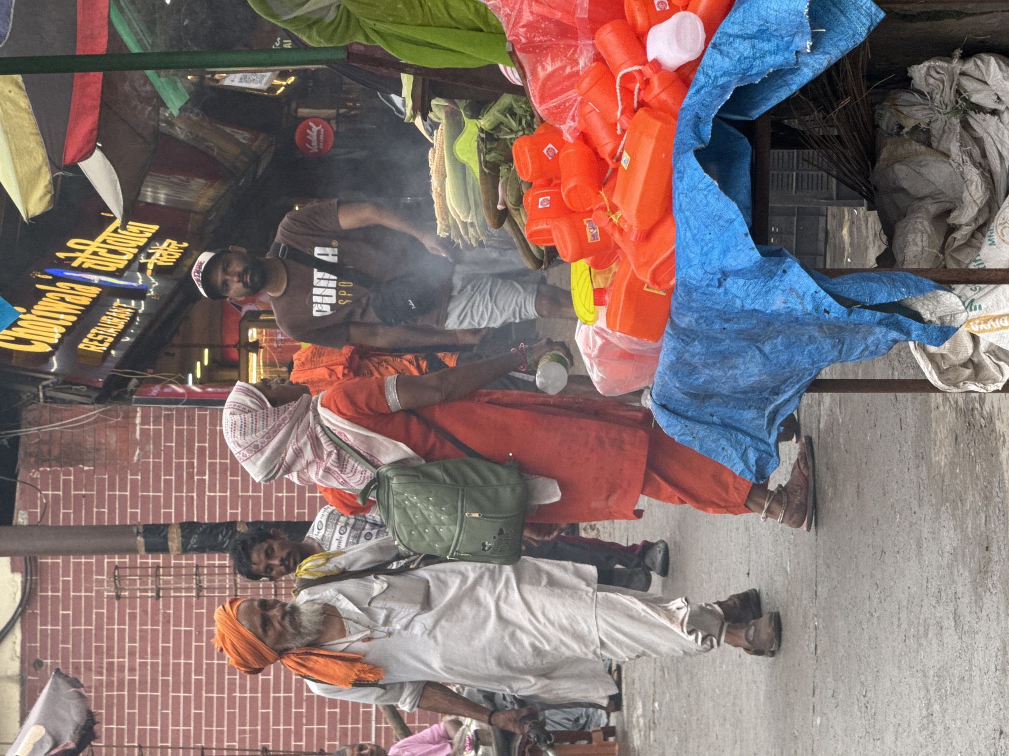 The image depicts a street scene in Rishikesh, India, likely near a market or food stall. In the foreground, a man wearing an orange turban and a white kurta walks with a long beard and a serious expression. To his right, a woman in an orange outfit and a white and pink headscarf carries a green backpack. Behind them, other individuals are visible, including a man in a brown t-shirt and a baseball cap. In the background, a sign for "Chotiwala Restaurant" is prominently displayed, suggesting a commercial area. The street appears to be paved and slightly wet. There are various objects on display, including a pile of bright orange plastic containers, bundles of corn, and other produce, along with some bundled sacks and palm fronds. The overall atmosphere suggests a bustling, possibly humid, day in a popular Indian city.