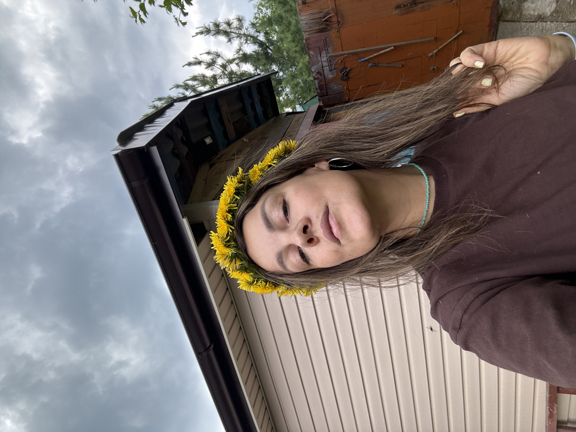 The image shows a woman with a dandelion crown on her head, looking at the camera. She is wearing a dark brown t-shirt and a light blue beaded necklace. Her hair is long and brown, and she is touching a lock of it with her right hand. She has a silver hoop earring in her left ear. In the background, there is a beige-sided building with a dark brown gutter. To the right of the building, there is a wooden fence and some trees. The sky is cloudy and gray. The scene appears to be outdoors, possibly in a rural area or a backyard.