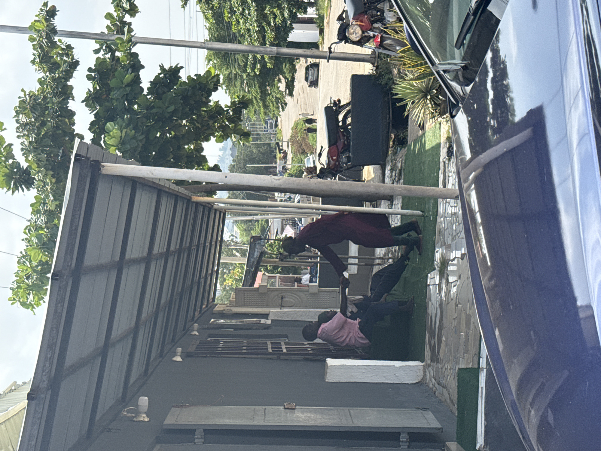 Two people are visible in the foreground, one wearing a maroon garment and the other in a pink shirt. They appear to be engaged in some physical activity, possibly a form of training or exercise, on a paved area. Behind them, a dark-colored vehicle is parked, and further in the background, there are trees, buildings, and other vehicles on a road. The scene suggests an outdoor setting, possibly in a residential or commercial area.
The lighting indicates it is daytime, and the sky is overcast. The overall atmosphere is casual. The people's body language suggests focus and exertion. There are no discernible signs or text in the visible portion of the image. The presence of lush greenery and the style of buildings could be indicative of a tropical or subtropical climate, consistent with the provided location context of Abuja, Nigeria. The scene appears to be a candid snapshot of everyday life.