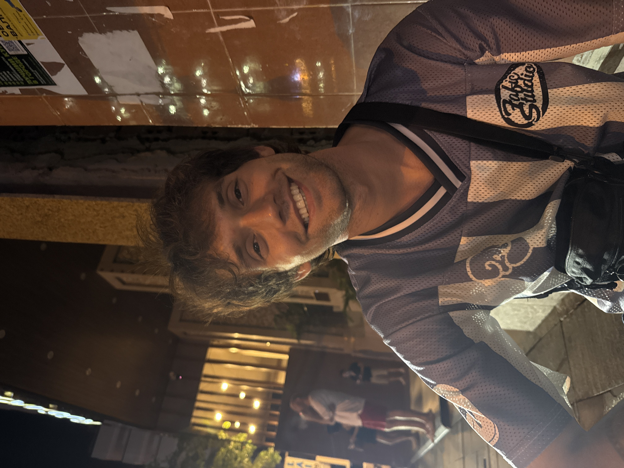 A man smiles at the camera while standing on a city street at night. He wears a patterned jersey with "Fabric Studio" on the chest and a black cross-body bag. The street is paved, and the background shows a building with lit windows and vegetation. People are visible in the distance. The location is likely in Nha Trang Ward, Vietnam, based on image metadata.