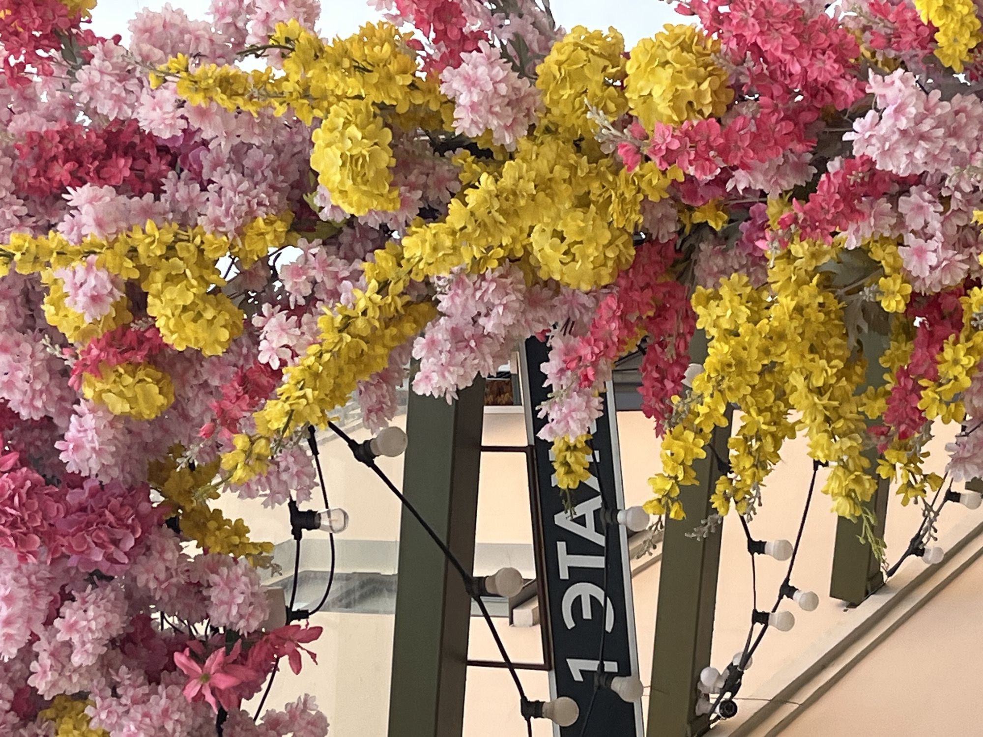 The image depicts a street scene in Izhevsk, Russia, identified by the Cyrillic text "1 ЭТАЖ" (1st floor) on a sign. The foreground is filled with dense artificial flowers in pink, red, and yellow, creating a canopy effect. Behind the floral display is an architectural structure, possibly part of a building facade or street decoration, with dark green supports. String lights with small, round bulbs are strung across the structure, adding to the festive ambiance. The background is out of focus but suggests a light-colored building exterior. There is no specific indication of a particular event or activity, but the decorations suggest a celebration or decorative installation. No people are visible in the image.