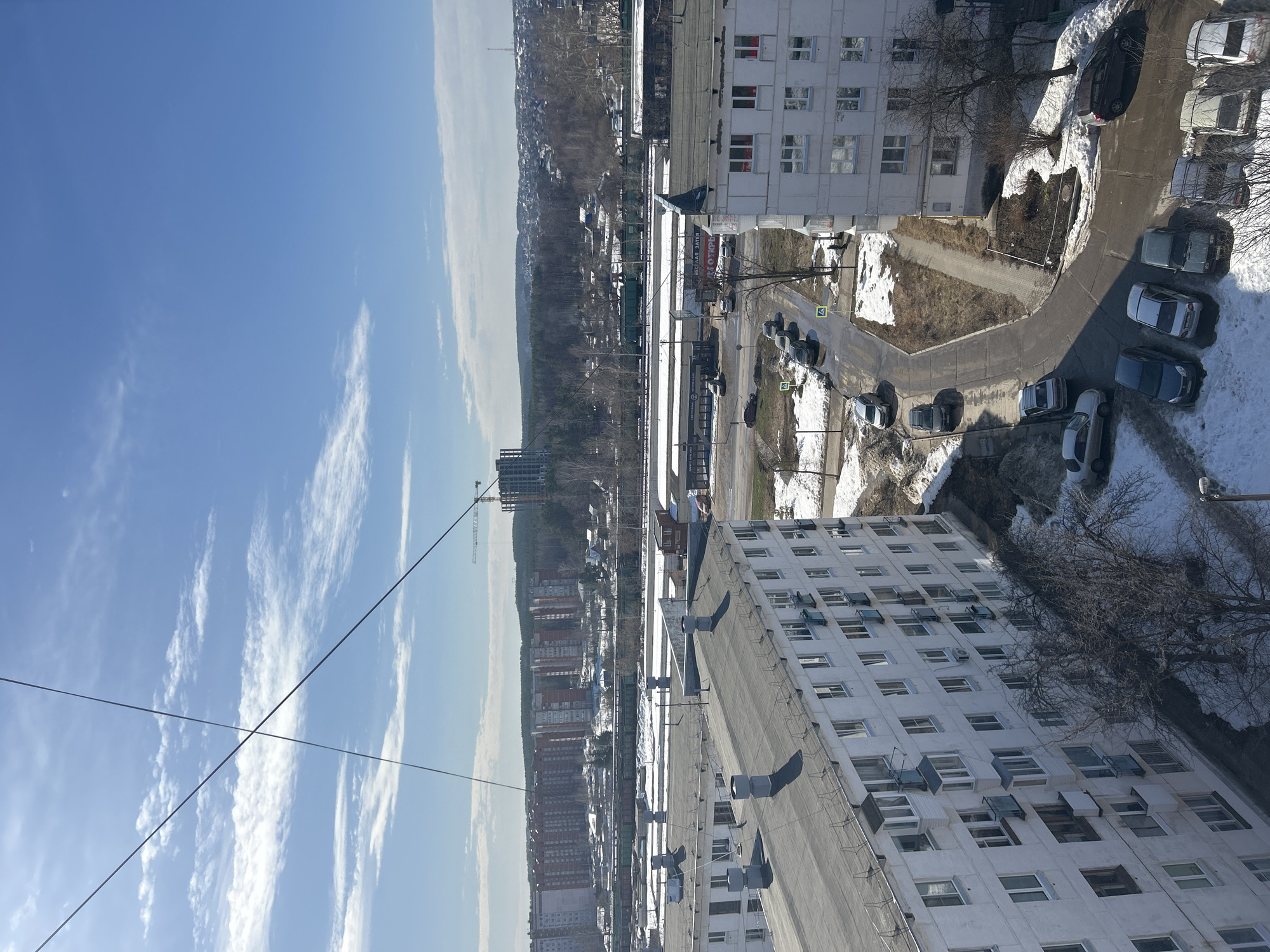 An elevated view captures a late winter or early spring urban landscape in Chaikovsky, Russia, under a bright blue sky with scattered white clouds. Two dark utility cables cross in the upper left foreground. In the mid-ground, two multi-story white residential buildings are prominent. The building on the left features a flat dark grey roof with multiple cylindrical vents and barbed wire along its edge; numerous windows and some enclosed balconies or air conditioning units are visible on its facade. A winding asphalt road separates these buildings, featuring approximately 15-20 parked cars of various models and colors (e.g., dark grey, silver, white, maroon) along its length and in adjacent parking areas. Yellow pedestrian crossing signs are visible on the road surface. Patches of residual snow, bare earth, and dormant deciduous trees are present around the buildings and roadways. A street light with an attached security camera is visible in the lower right.
Further into the background, a series of single-story commercial or industrial buildings with flat roofs and signs, including one that appears to read "ОТКРЫТ" (OPEN), border a railway line. Several green freight train cars are visible on these tracks. Beyond the railway, a dense urban residential area extends to the horizon, characterized by numerous multi-story apartment buildings, some with red brick sections, interspersed with bare trees and wooded hills. A tall construction crane is prominent on the right, positioned over a partially constructed high-rise building. A communication tower is visible on a distant hill to the right. No significant human activity or direct interactions are clearly identifiable within the scene.